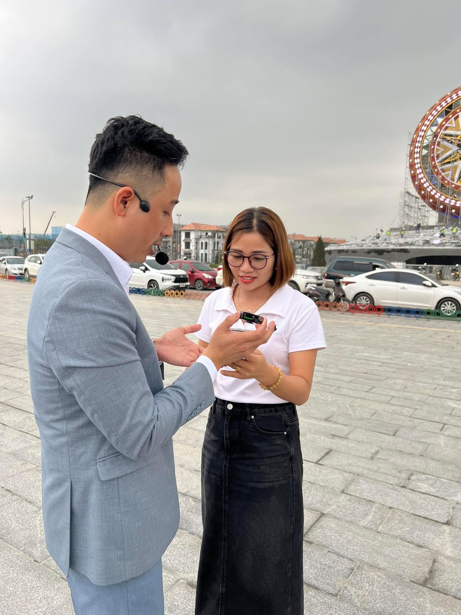 Đăng Dương mặc vest xám đeo micro cài tai, trao đổi cùng một nữ đồng nghiệp áo trắng ngoài quảng trường một dự án