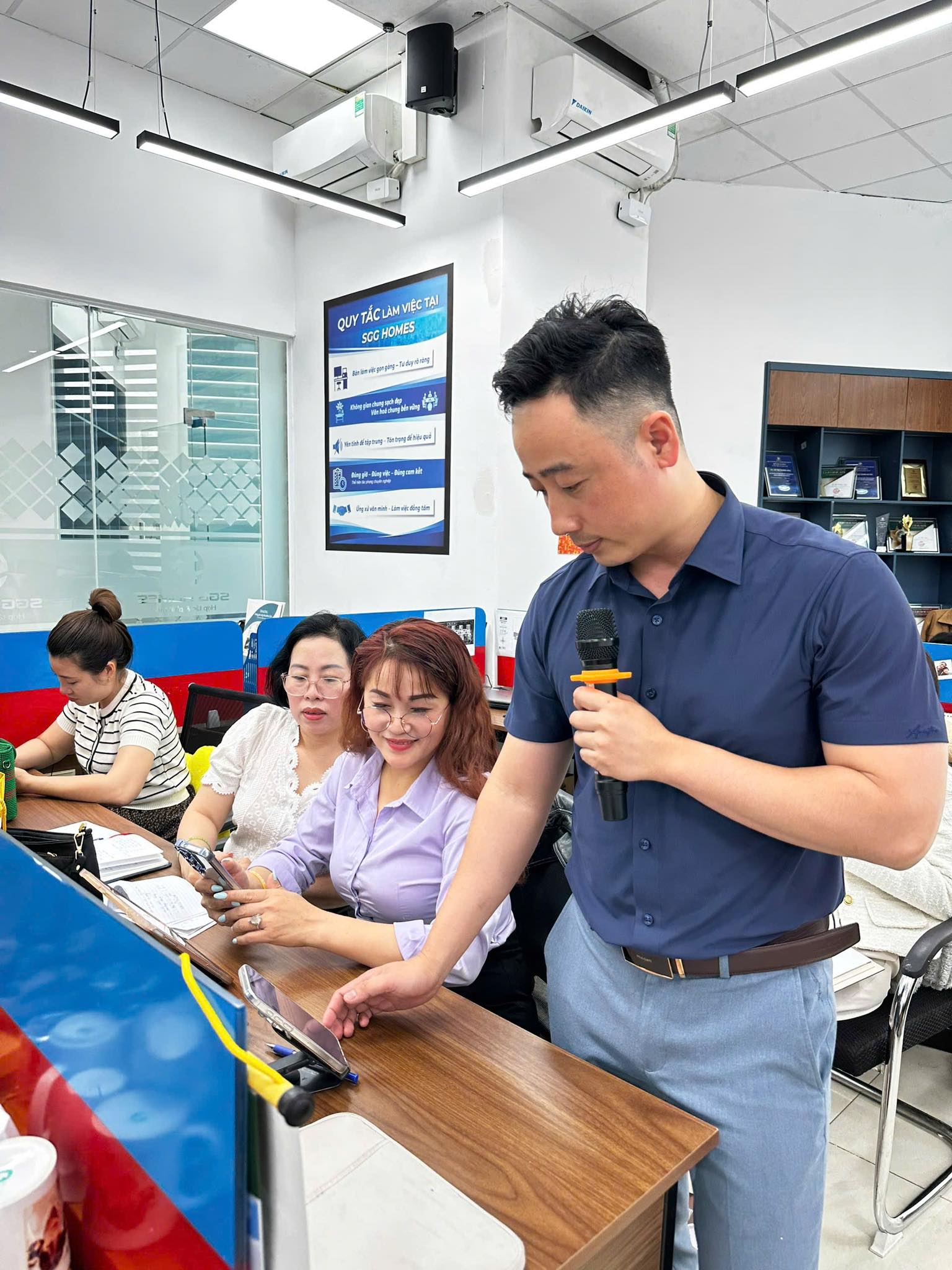 Đăng Dương cầm micro đứng cạnh học viên đang thao tác trên điện thoại trong lớp học