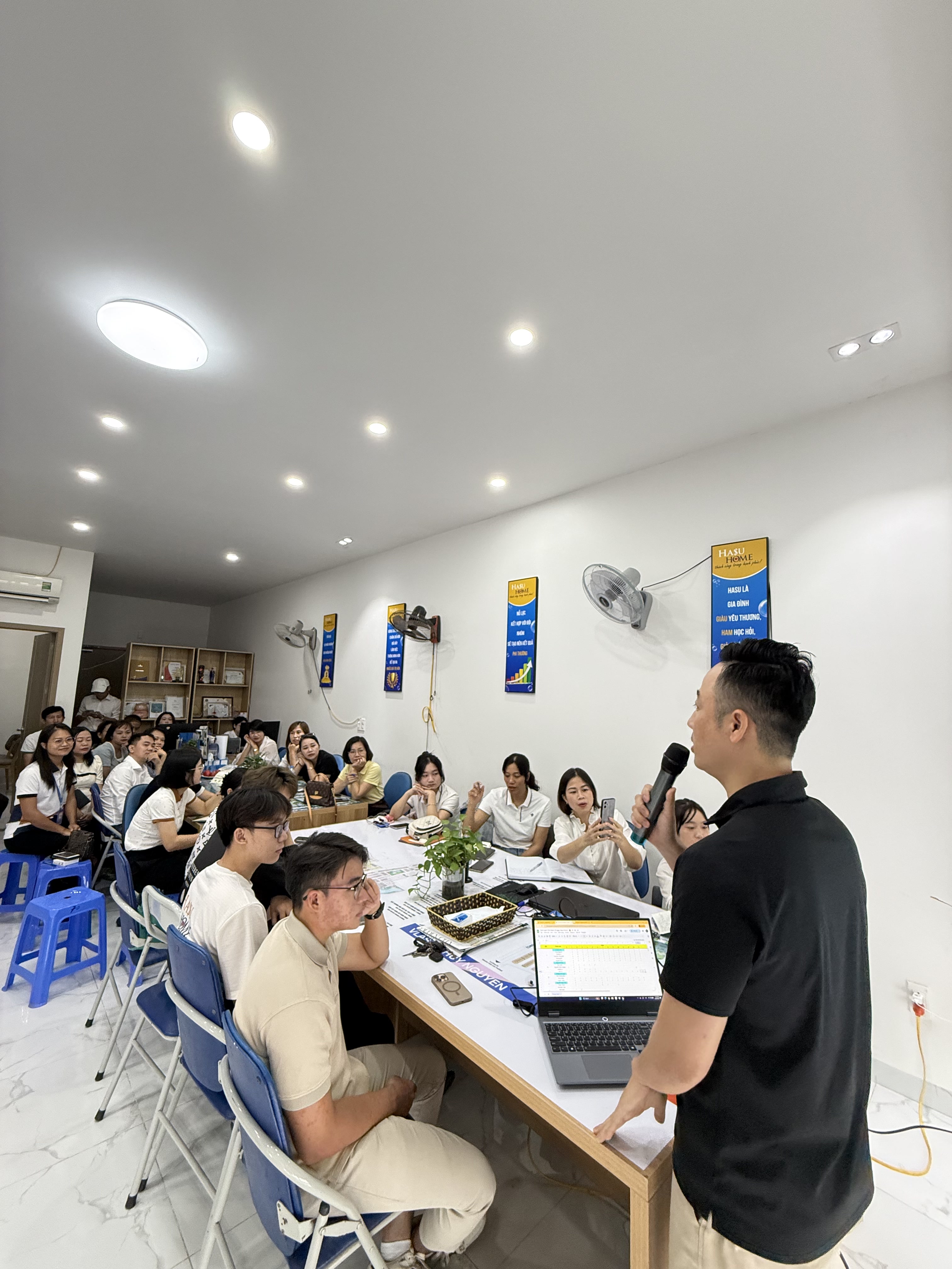 Đăng Dương áo polo đen cầm micro đứng cạnh bàn học viên trong phòng đào tạo có băng rôn xanh dương