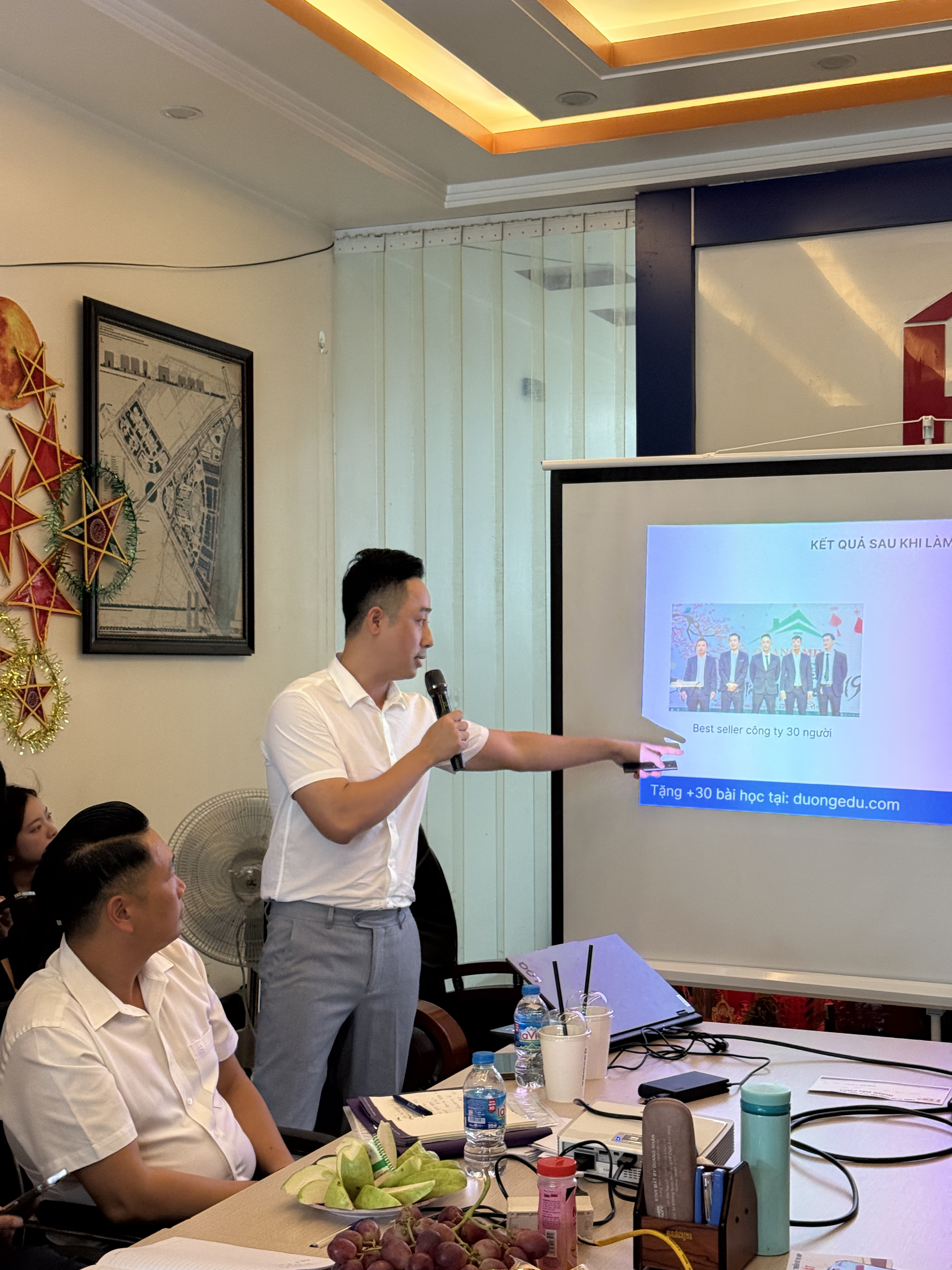 Đăng Dương mặc áo polo trắng cầm micro, tay phải chỉ vào màn chiếu đang hiện ảnh một nhóm người cùng dòng tiêu đề về kết quả đạt được sau khi bắt tay vào làm