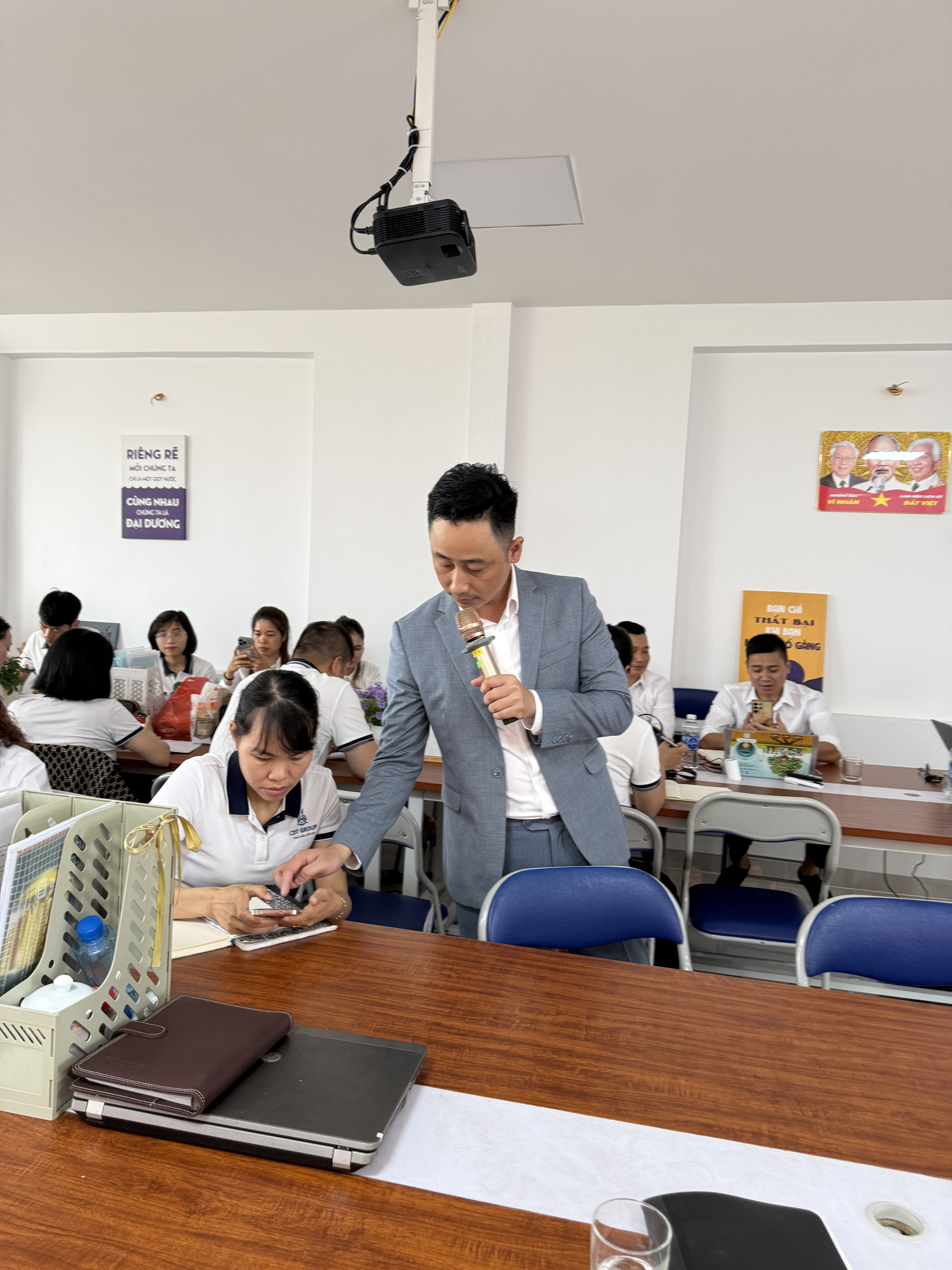 Đăng Dương mặc vest xám cầm micro, nghiêng người cúi xuống nhìn chiếc điện thoại trên tay một học viên nữ đang ngồi, trước mặt cô là quyển sổ đang ghi dở