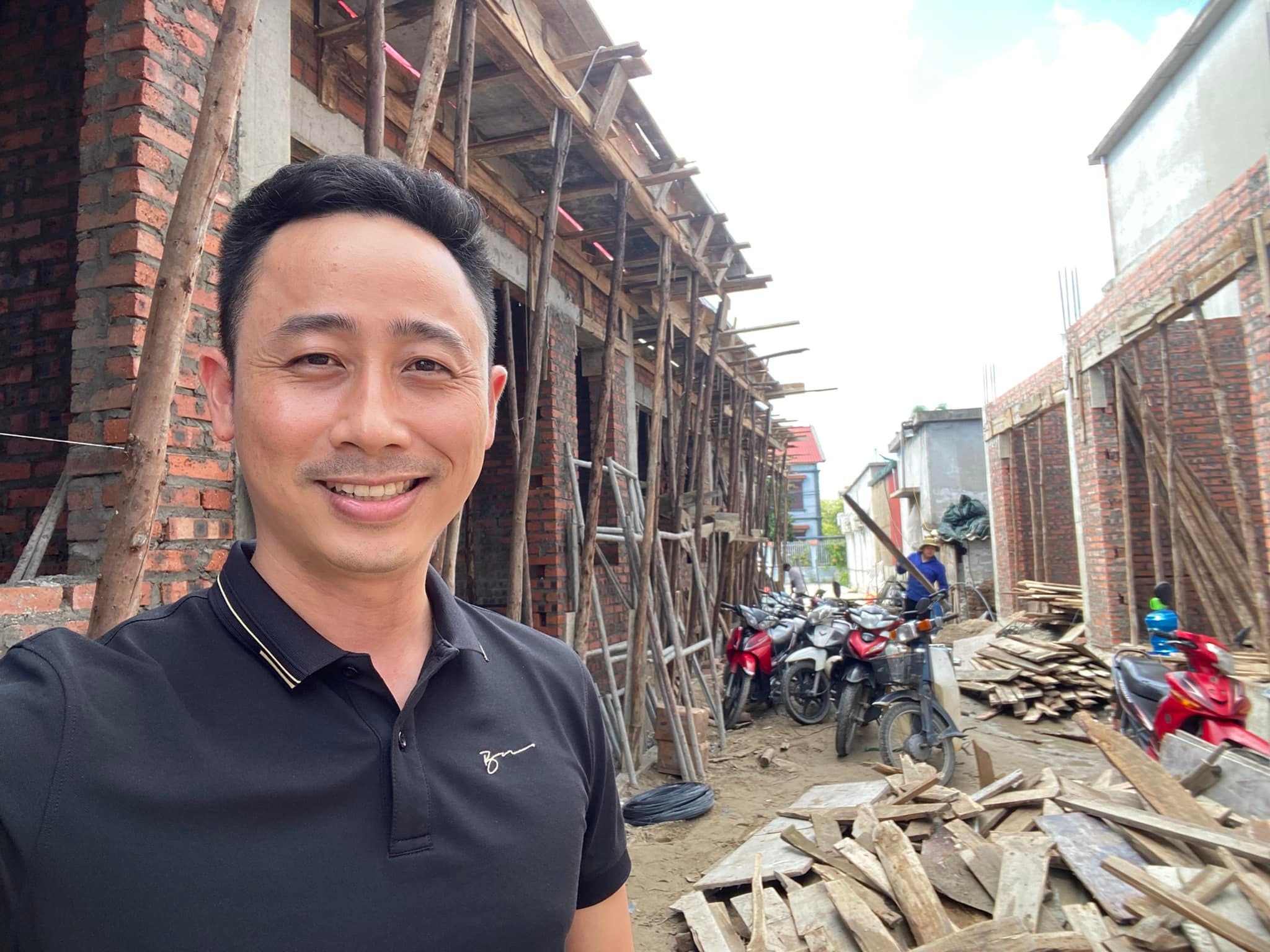 Đăng Dương chụp ảnh selfie trước một dãy nhà đang xây thô với giàn giáo tre, thợ đang làm việc và xe máy dựng dọc lối đi