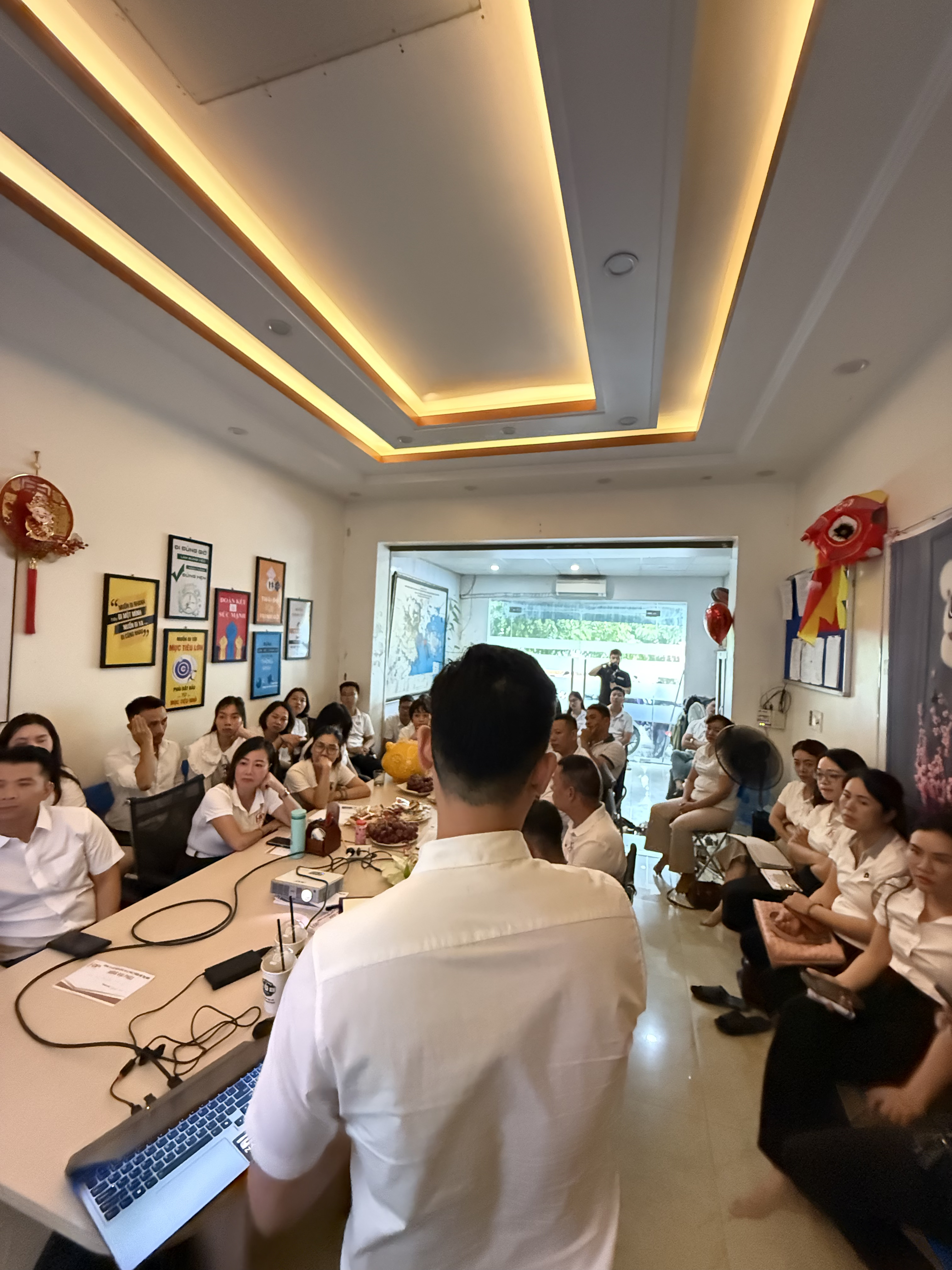 Đăng Dương mặc sơ mi trắng cầm micro đứng cạnh màn chiếu, khoảng tám người ngồi hai bên bàn với sổ ghi chép mở và bút trên tay
