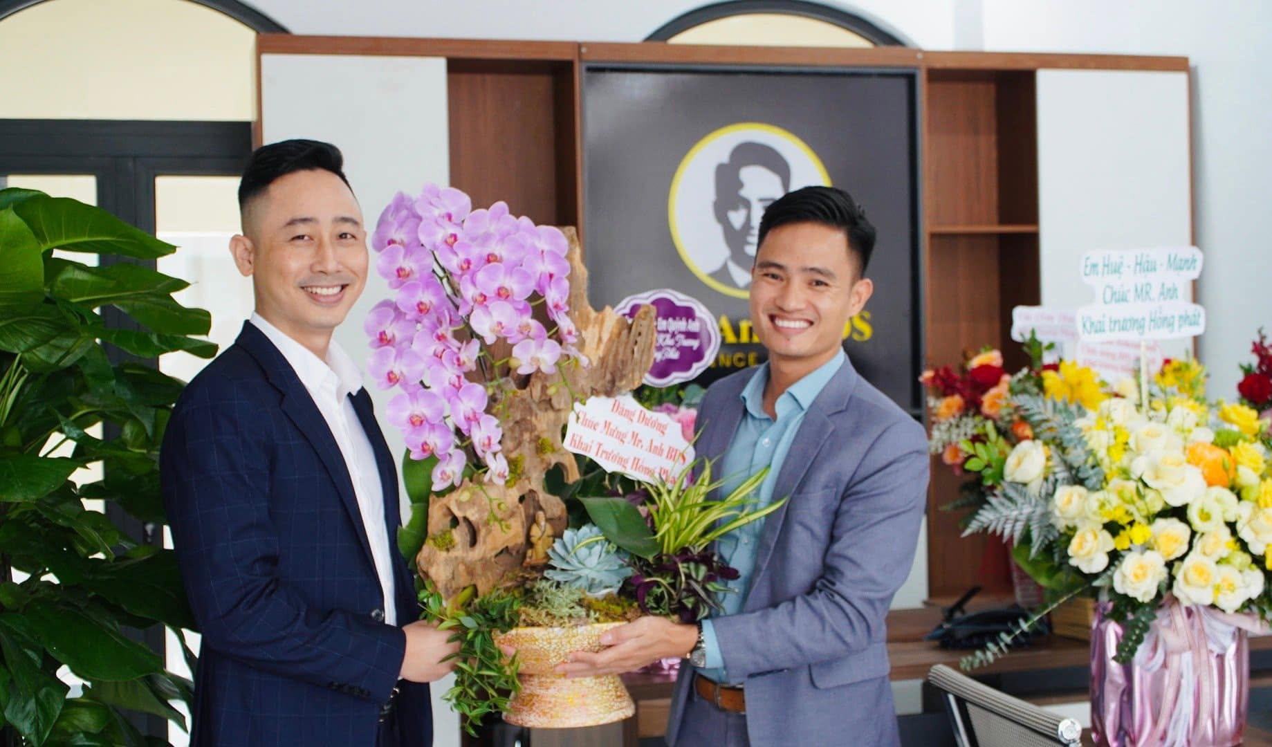 Đăng Dương và một học viên cùng cầm chậu lan tím trong lễ khai trương văn phòng bất động sản, xung quanh là hoa chúc mừng