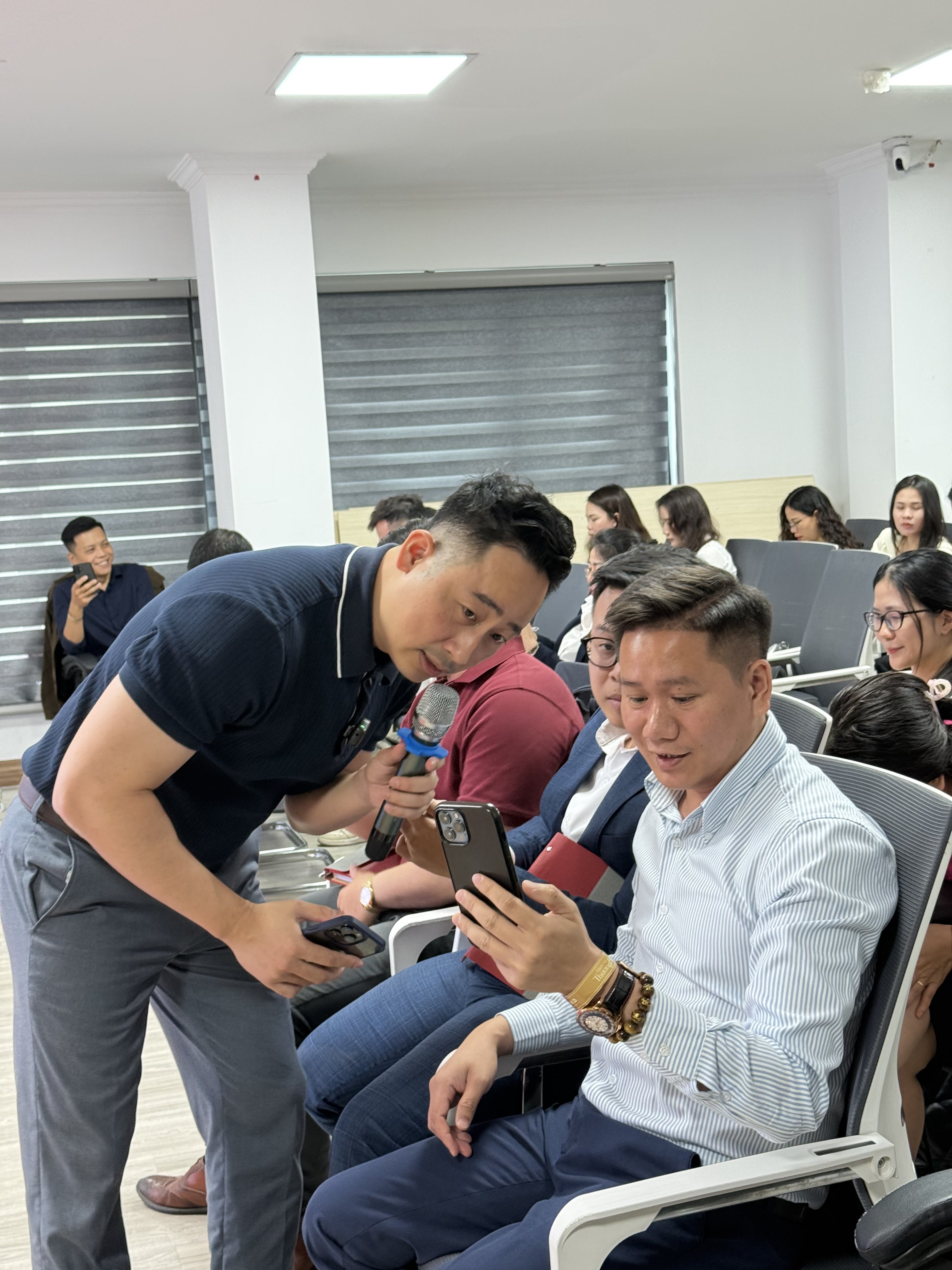 Đăng Dương mặc áo polo navy cúi người, một tay cầm micro một tay cầm điện thoại, đang hướng dẫn một môi giới mặc sơ mi kẻ sọc đang ngồi giơ điện thoại lên xem