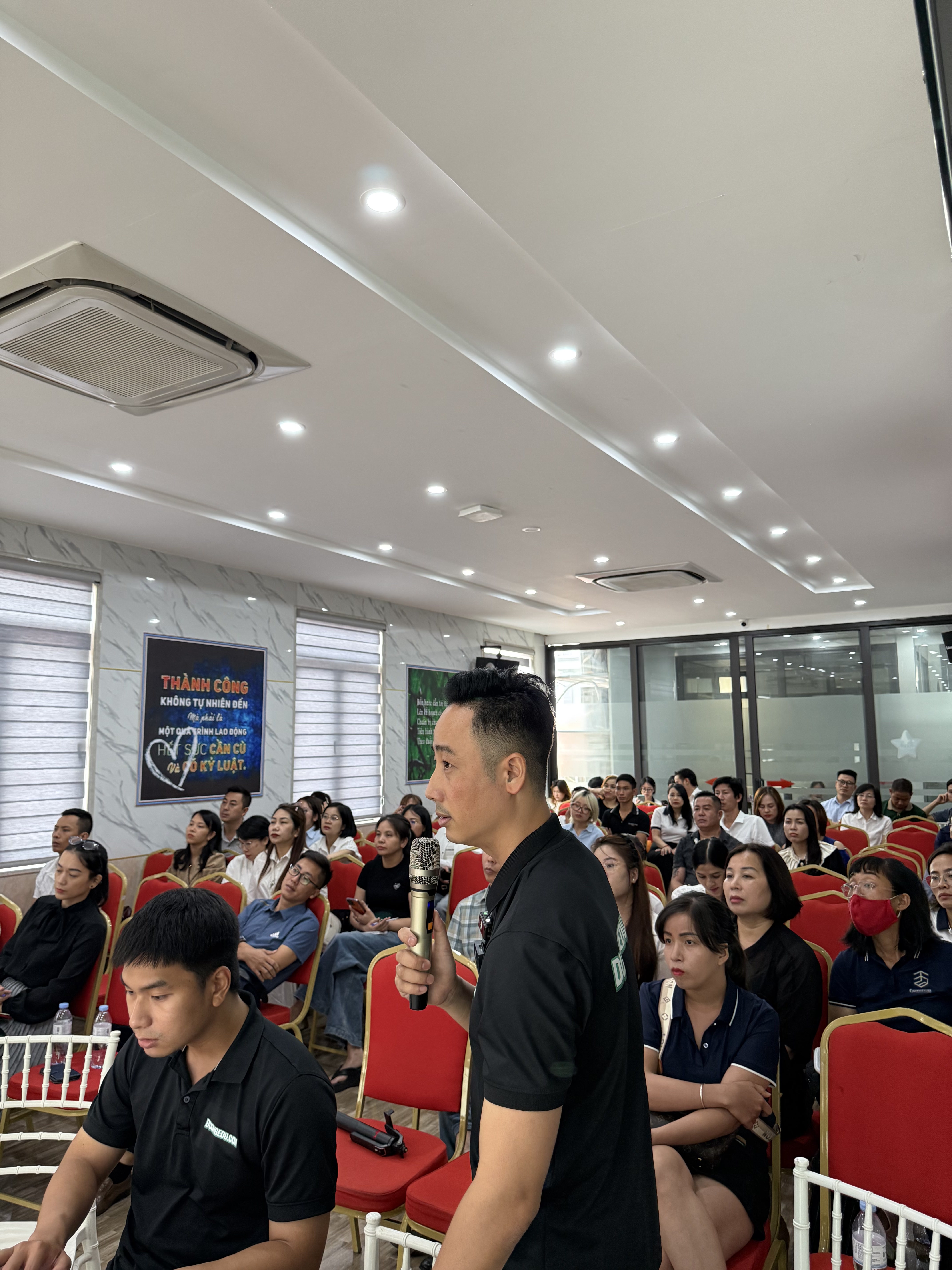 Đăng Dương mặc áo polo đen cầm micro đứng giữa hàng ghế nói với khoảng bốn năm chục người ngồi kín trên ghế bọc nhung đỏ
