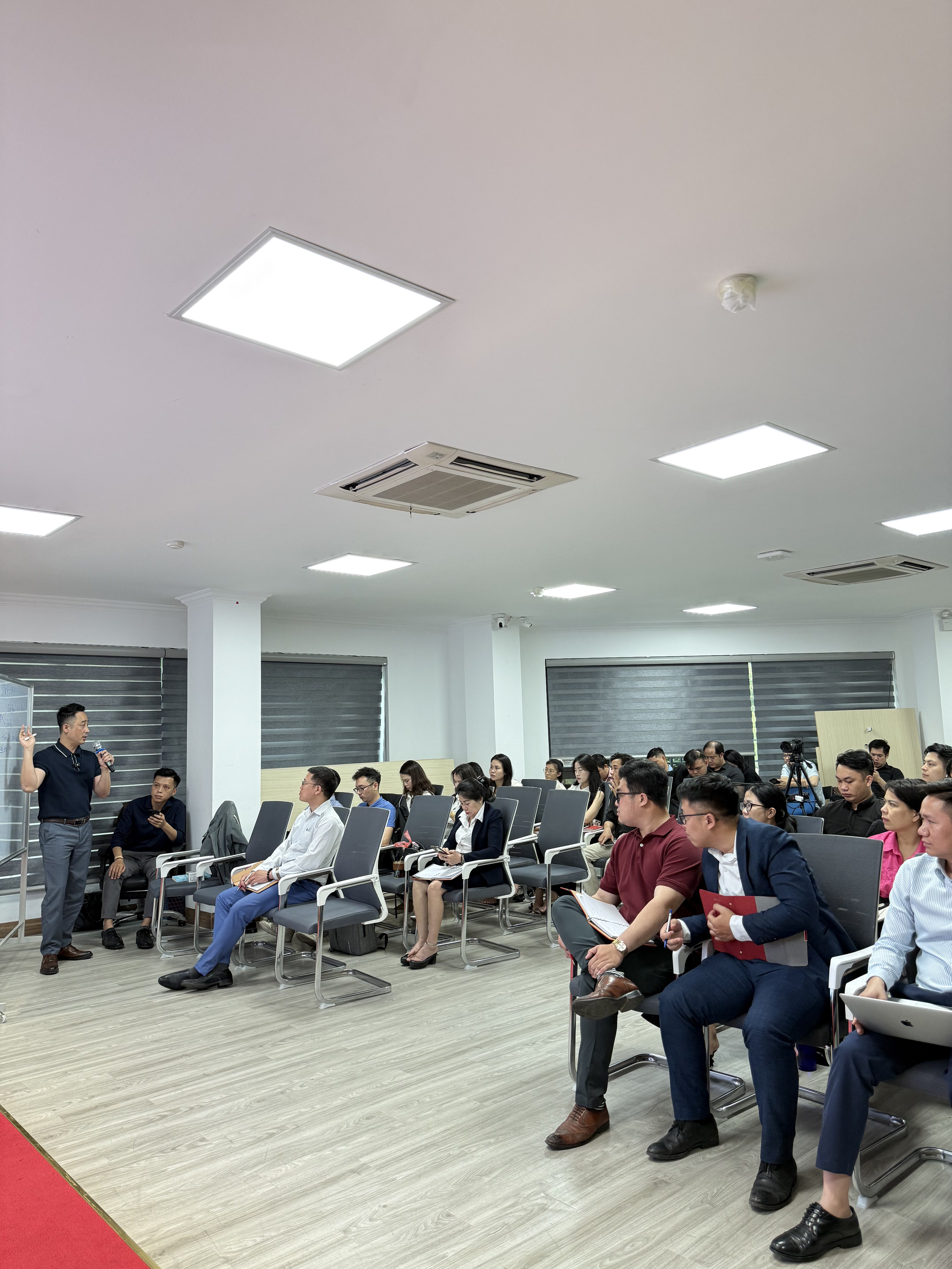 Khoảng ba mươi môi giới mặc sơ mi trắng và vest ngồi kín các hàng ghế xám, nhiều người cầm sổ và bút, một người mở laptop trên đùi, giảng viên đứng phía xa bên bảng trắng