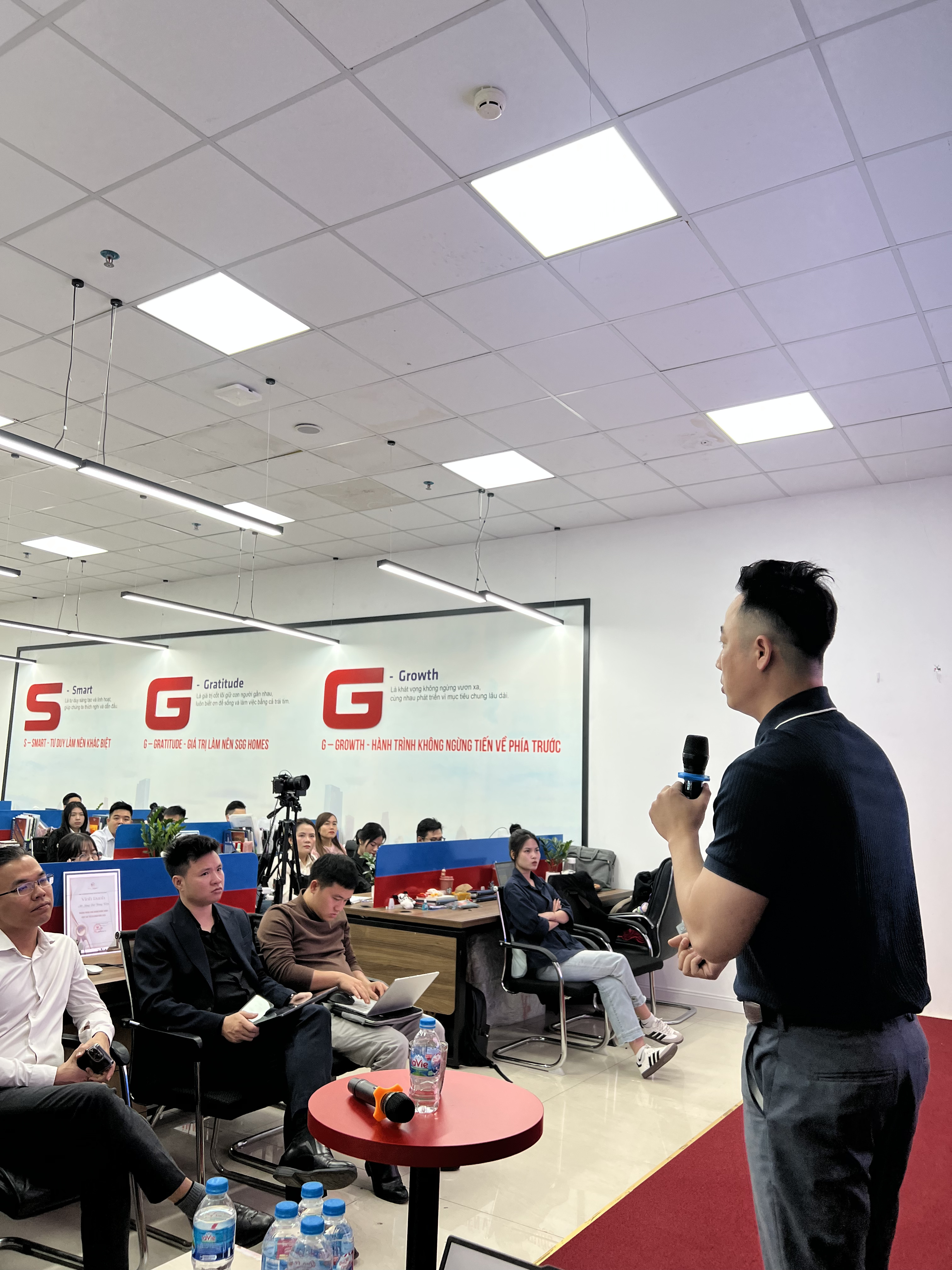 Đăng Dương mặc áo polo đen cầm micro đứng nói trước một sàn giao dịch, các môi giới ngồi nghe, một người cầm điện thoại, một người đang gõ laptop