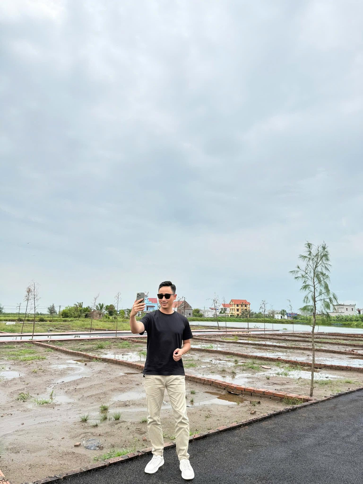 Một người đứng quay video bằng điện thoại tại khu đất nền đang san lấp, phía sau là các lô đất chia bằng gạch và nhà dân xa xa