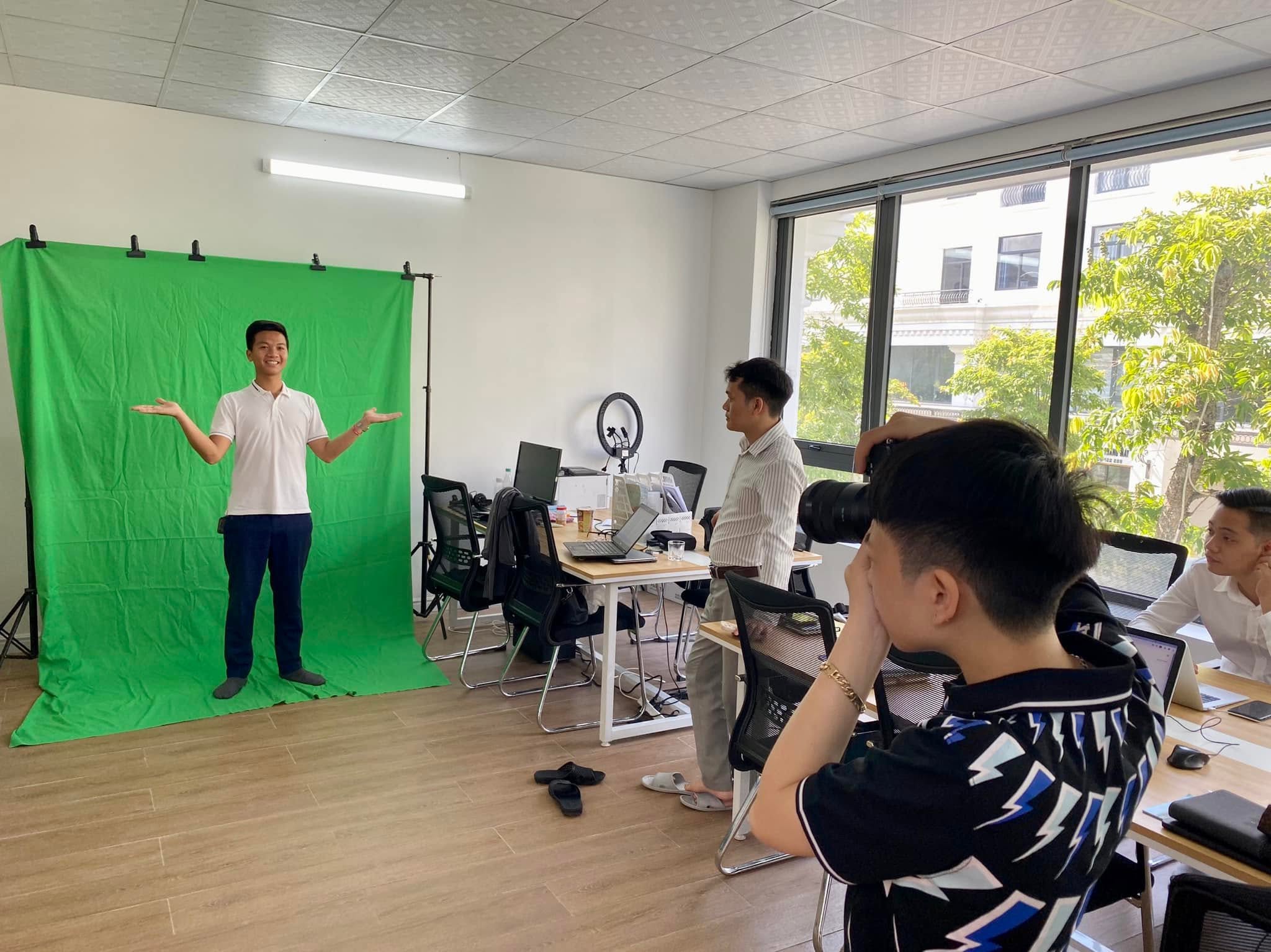 Một người đứng trước phông xanh trong văn phòng, người khác cầm máy ảnh ống tele chụp lại, bàn làm việc và laptop ở phía sau