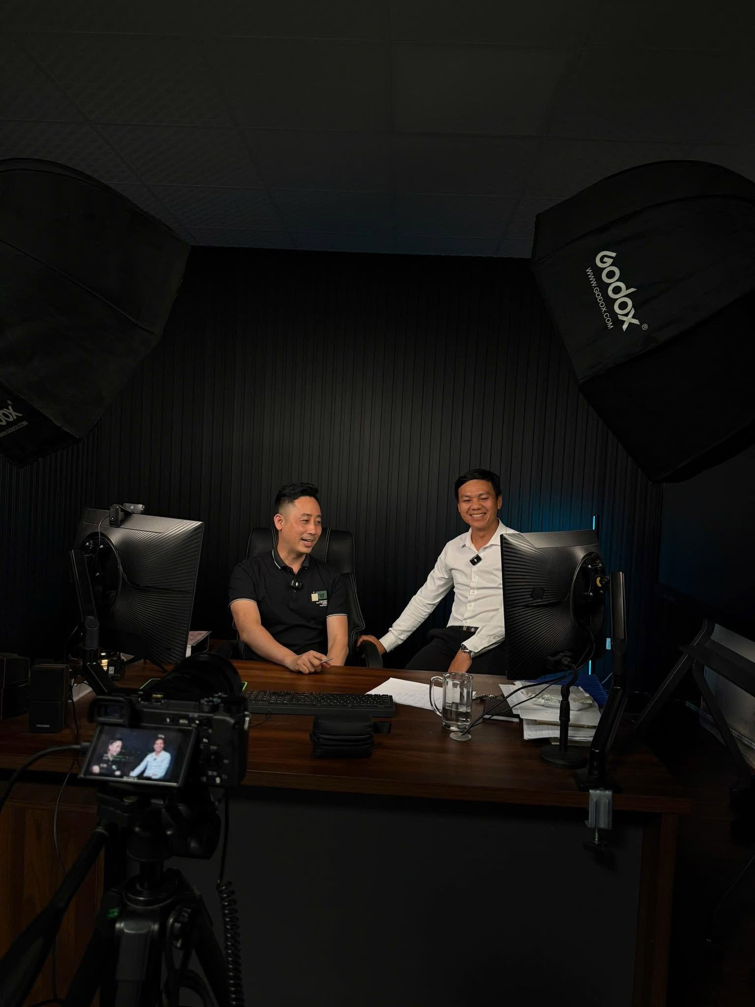 Hai người đàn ông ngồi trò chuyện trước máy quay trong phòng studio có đèn softbox, bàn làm việc và màn hình máy tính