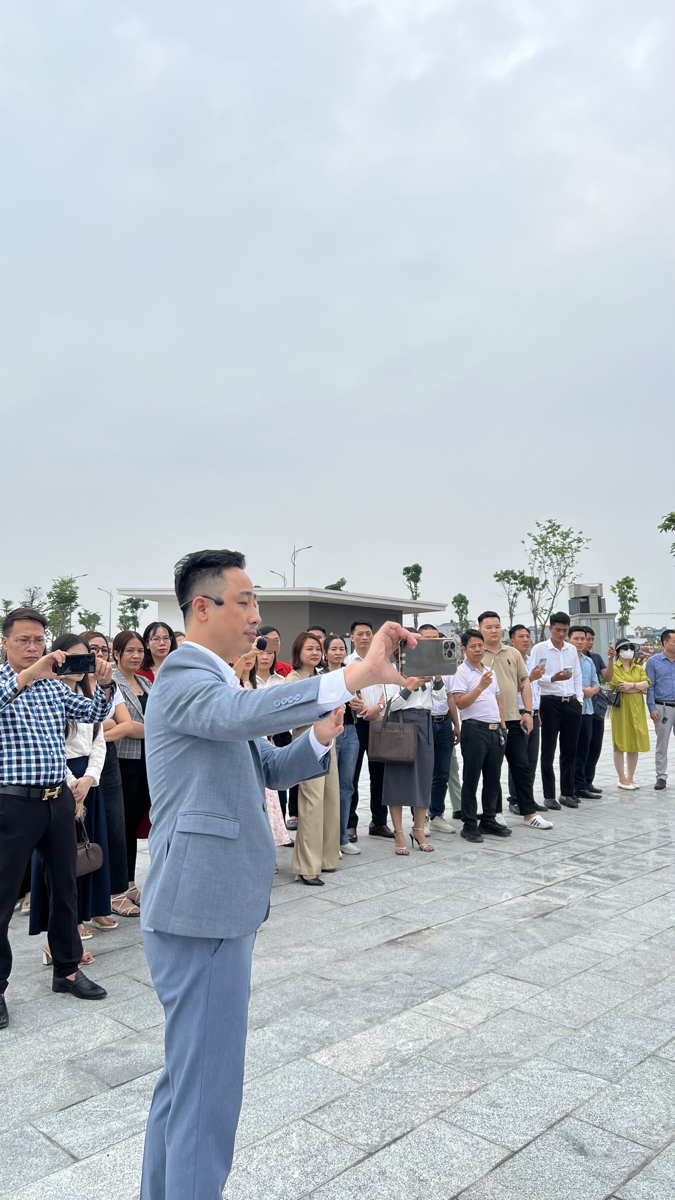 Đăng Dương mặc vest xanh xám đeo micro cài tai đứng giơ điện thoại ngang làm mẫu động tác quay, phía sau khoảng hai mươi môi giới đứng thành hàng cũng giơ điện thoại làm theo