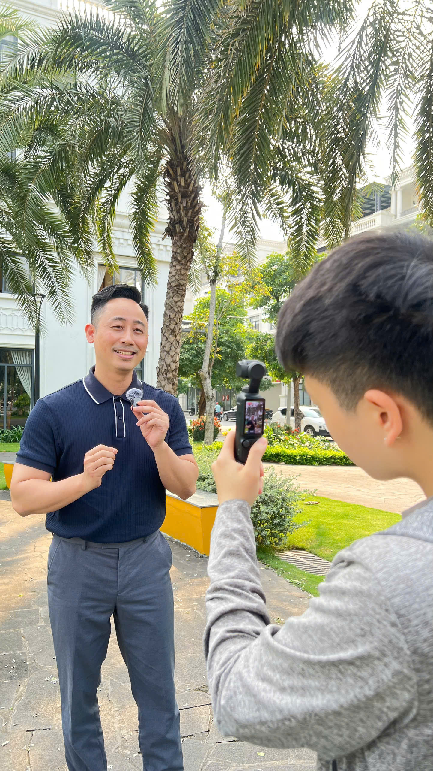 Đăng Dương mặc áo polo xanh navy đang cài micro không dây lên áo, bên cạnh là một người cầm máy quay nhỏ ghi hình, phía sau là dãy nhà trắng và hàng cọ