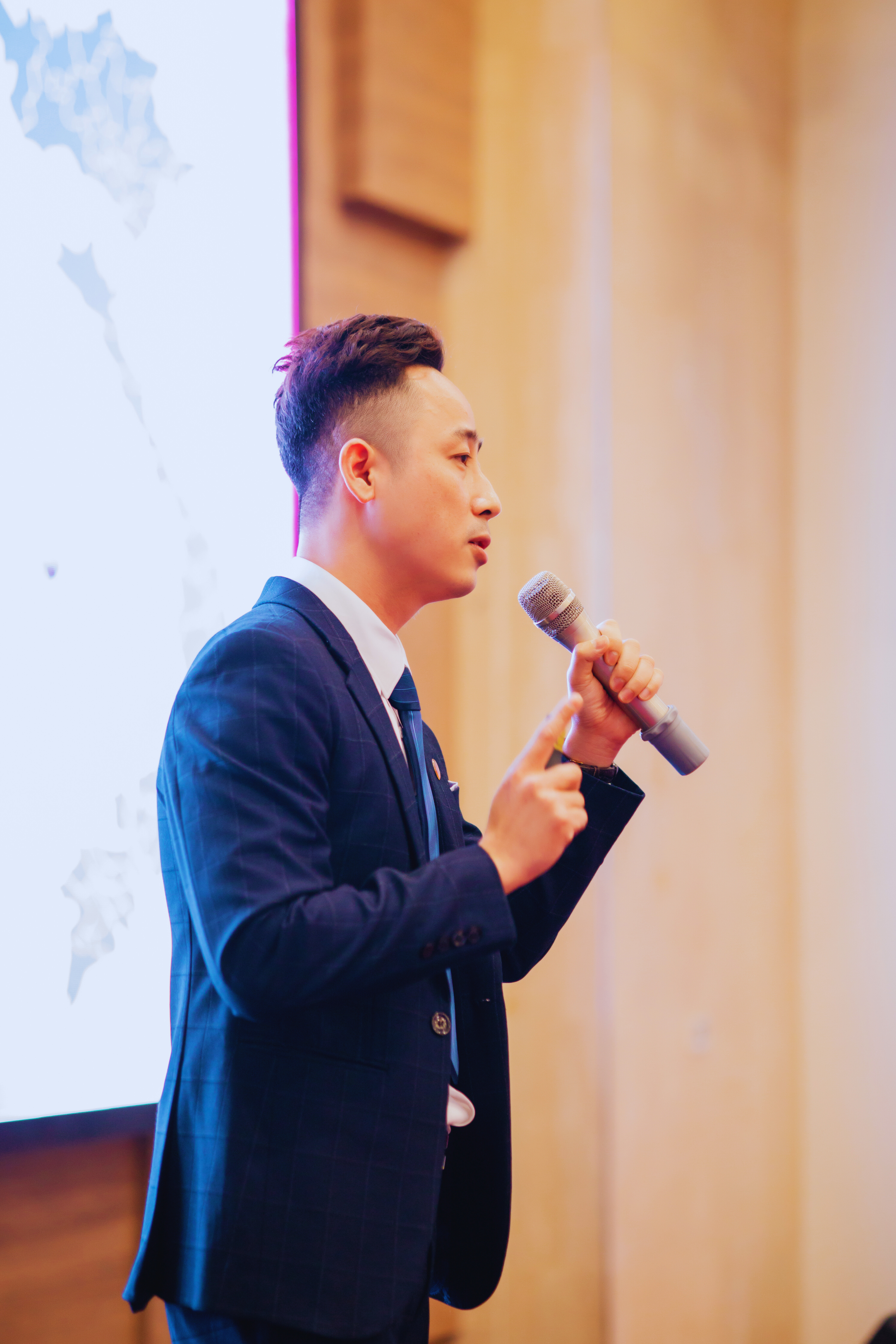 Đăng Dương mặc vest xanh than cầm micro nói trước màn chiếu lớn trong một hội trường