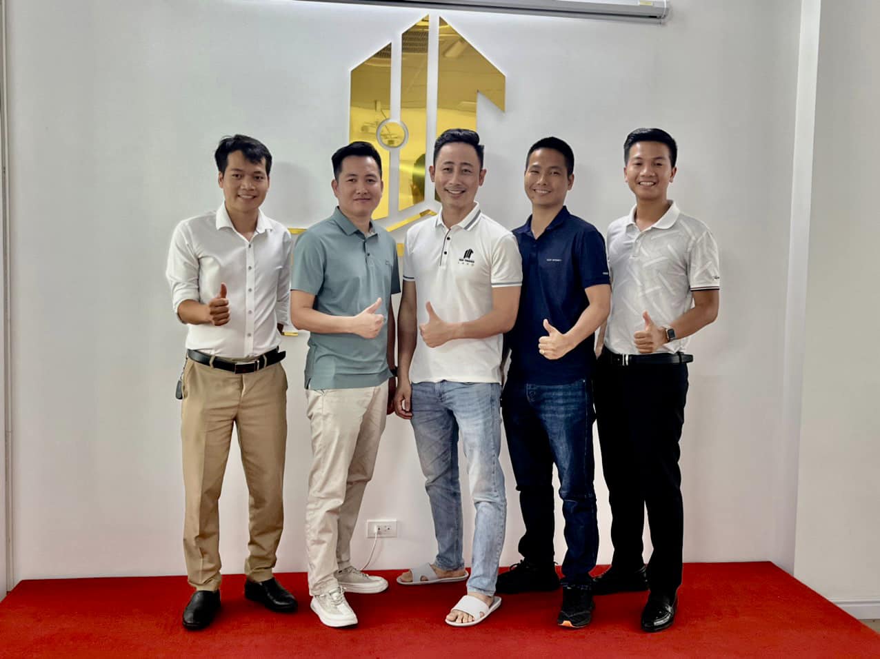 Năm người đứng giơ ngón tay cái trước bức tường có logo vàng của một sàn bất động sản, thảm đỏ dưới chân