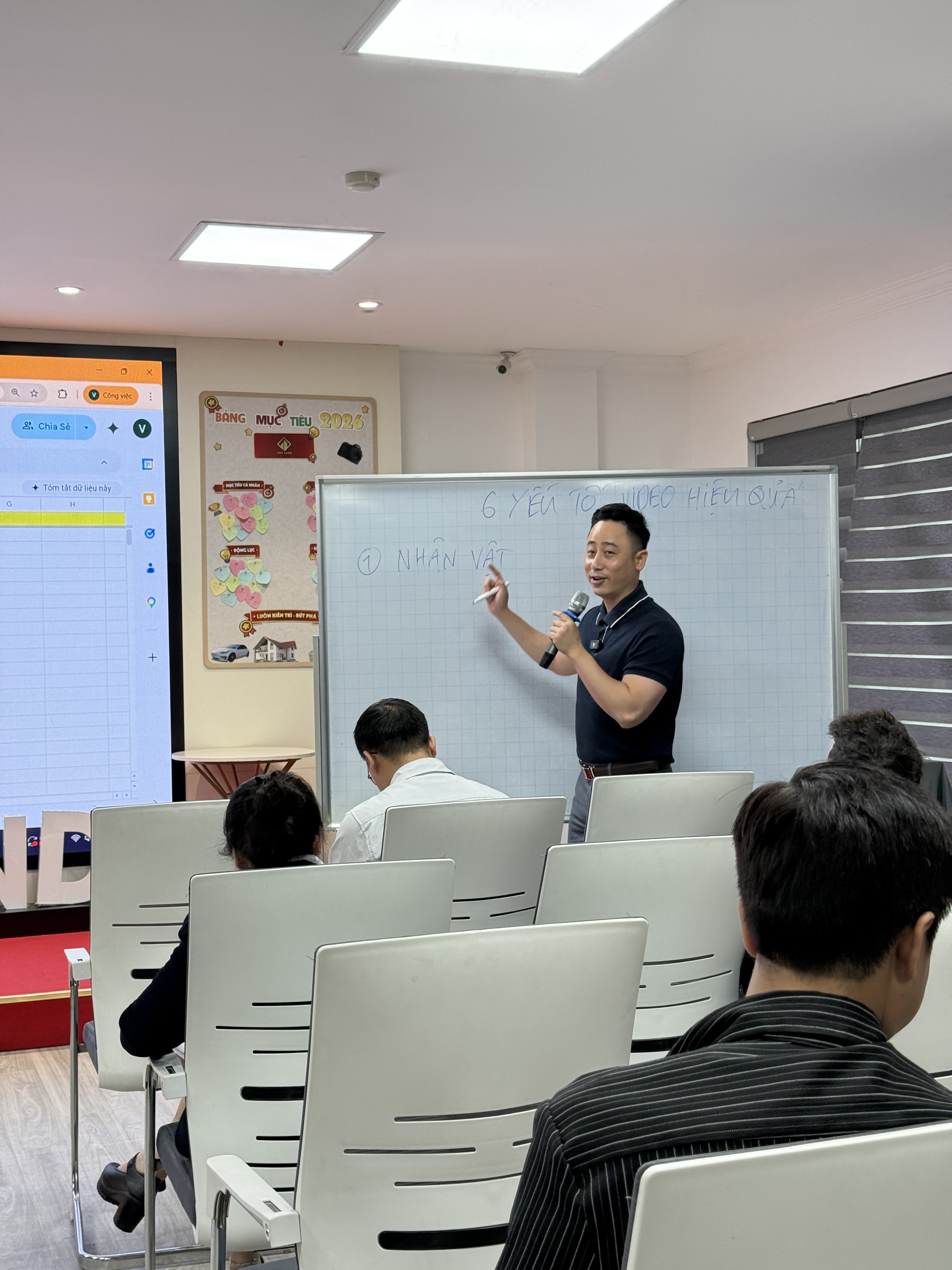 Đăng Dương mặc áo polo đen, một tay cầm micro, tay kia cầm bút dạ chỉ lên tấm bảng trắng đã viết sáu yếu tố của một video hiệu quả