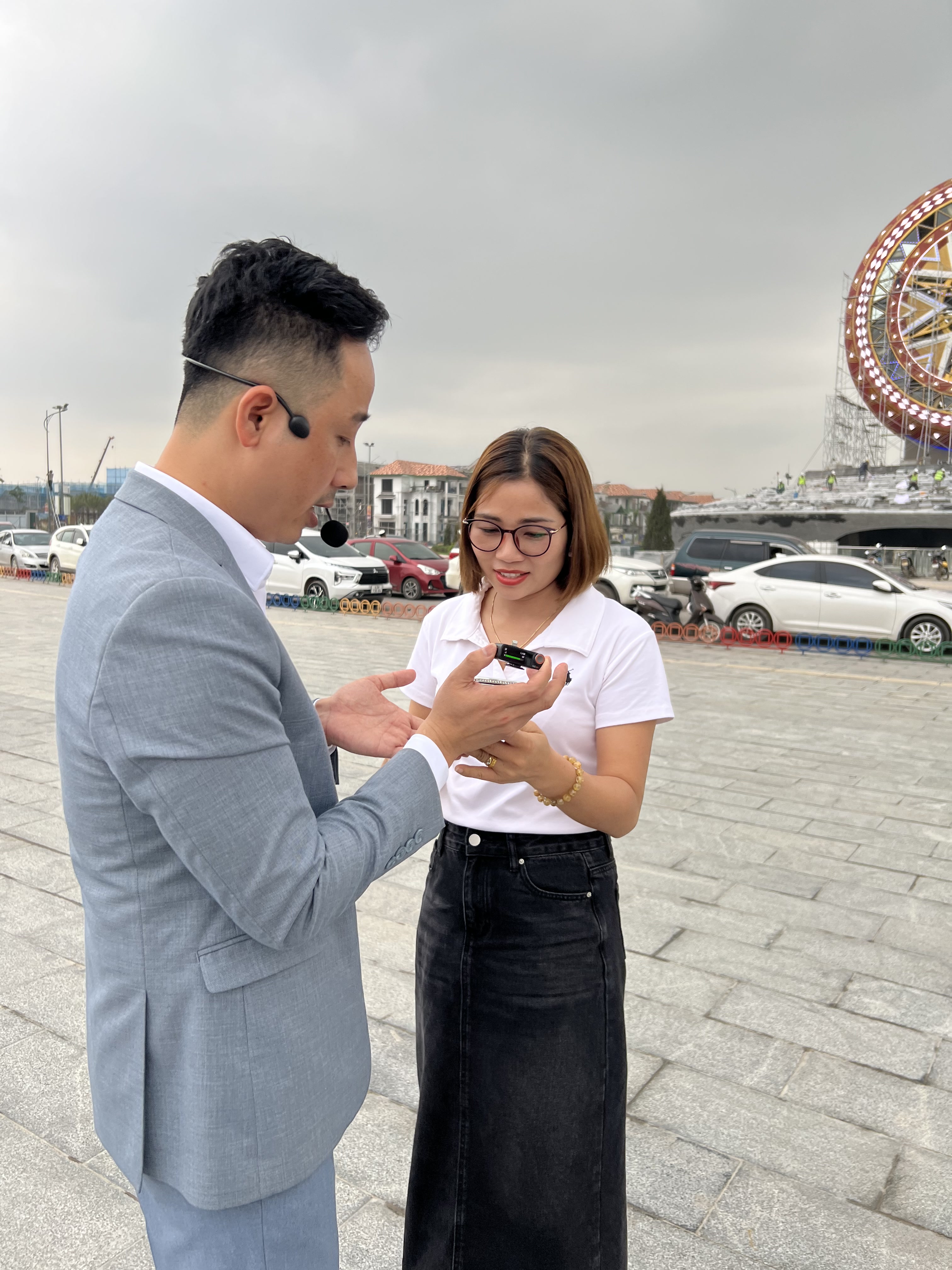 Đăng Dương mặc vest xám đeo micro cài tai đang hướng dẫn một nữ môi giới áo trắng cách gắn micro thu âm lên áo, phía sau là quảng trường một dự án đang thi công