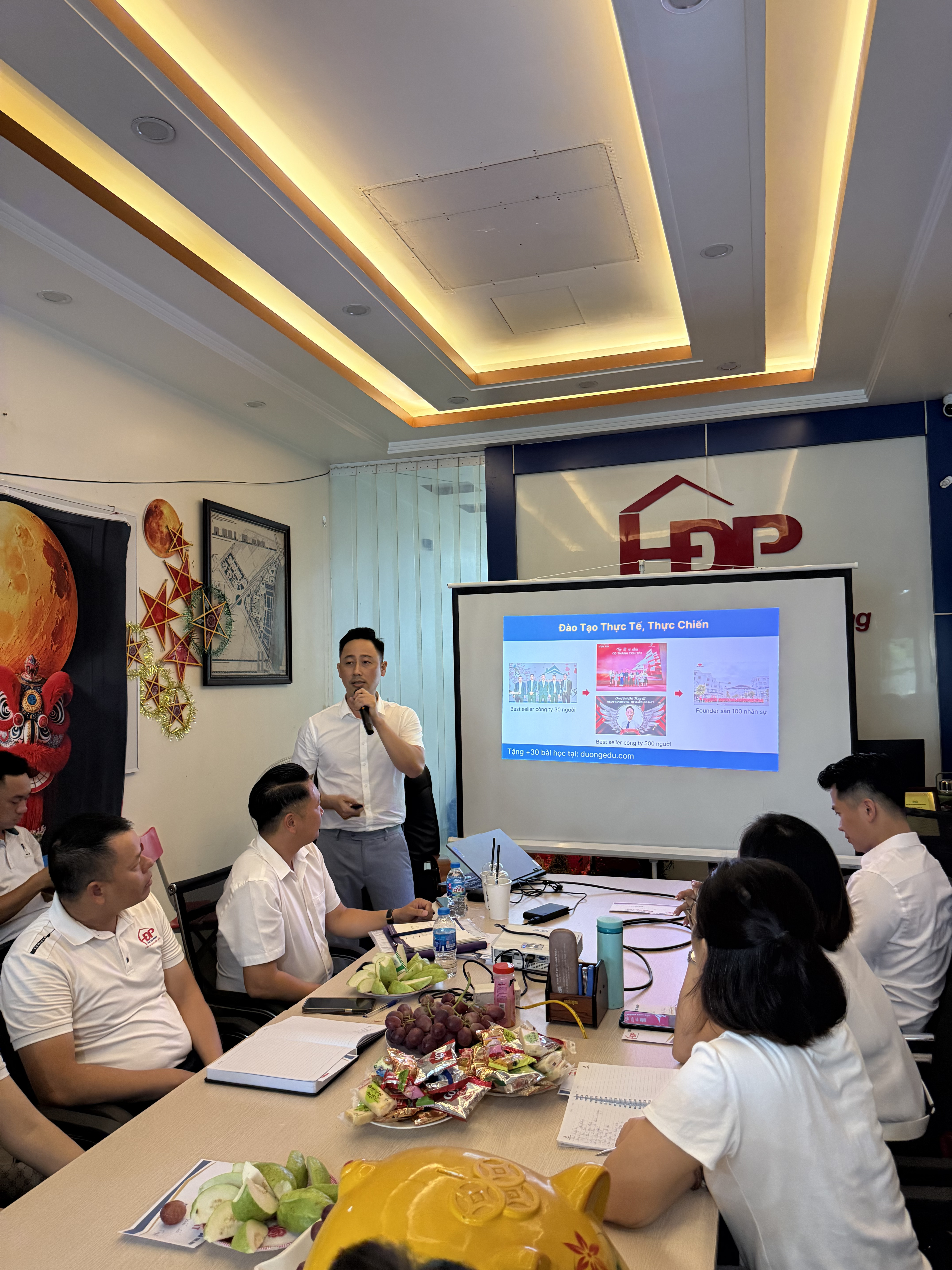 Đăng Dương mặc sơ mi trắng đứng giữa phòng họp đang nói, khoảng ba mươi người mặc đồng phục áo trắng ngồi kín quanh bàn dài và dọc hai bên tường cùng nhìn về phía anh