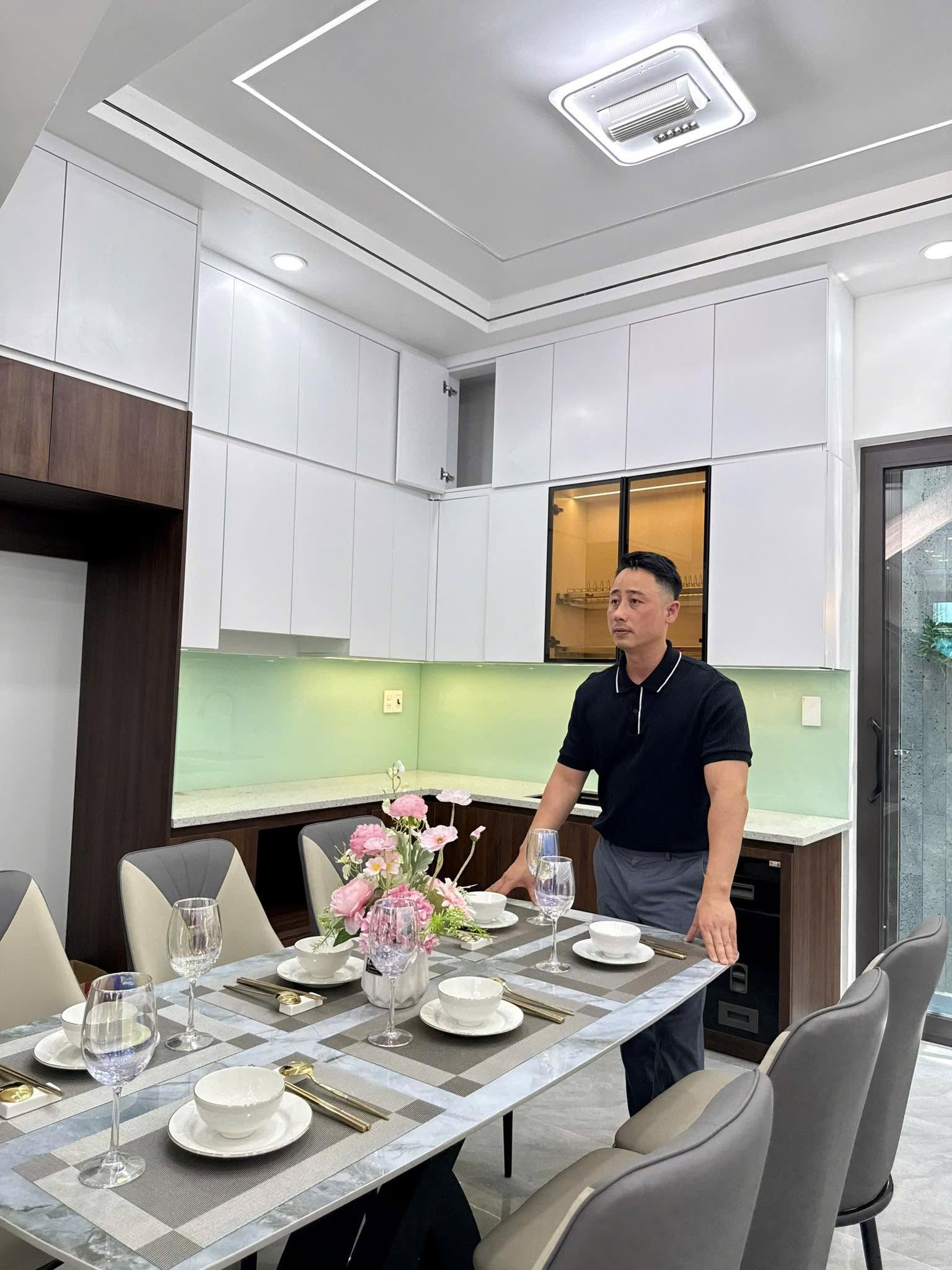 Đăng Dương đứng bên bàn ăn đã bày sẵn bát đĩa trong một căn nhà mới hoàn thiện, phía sau là tủ bếp trắng