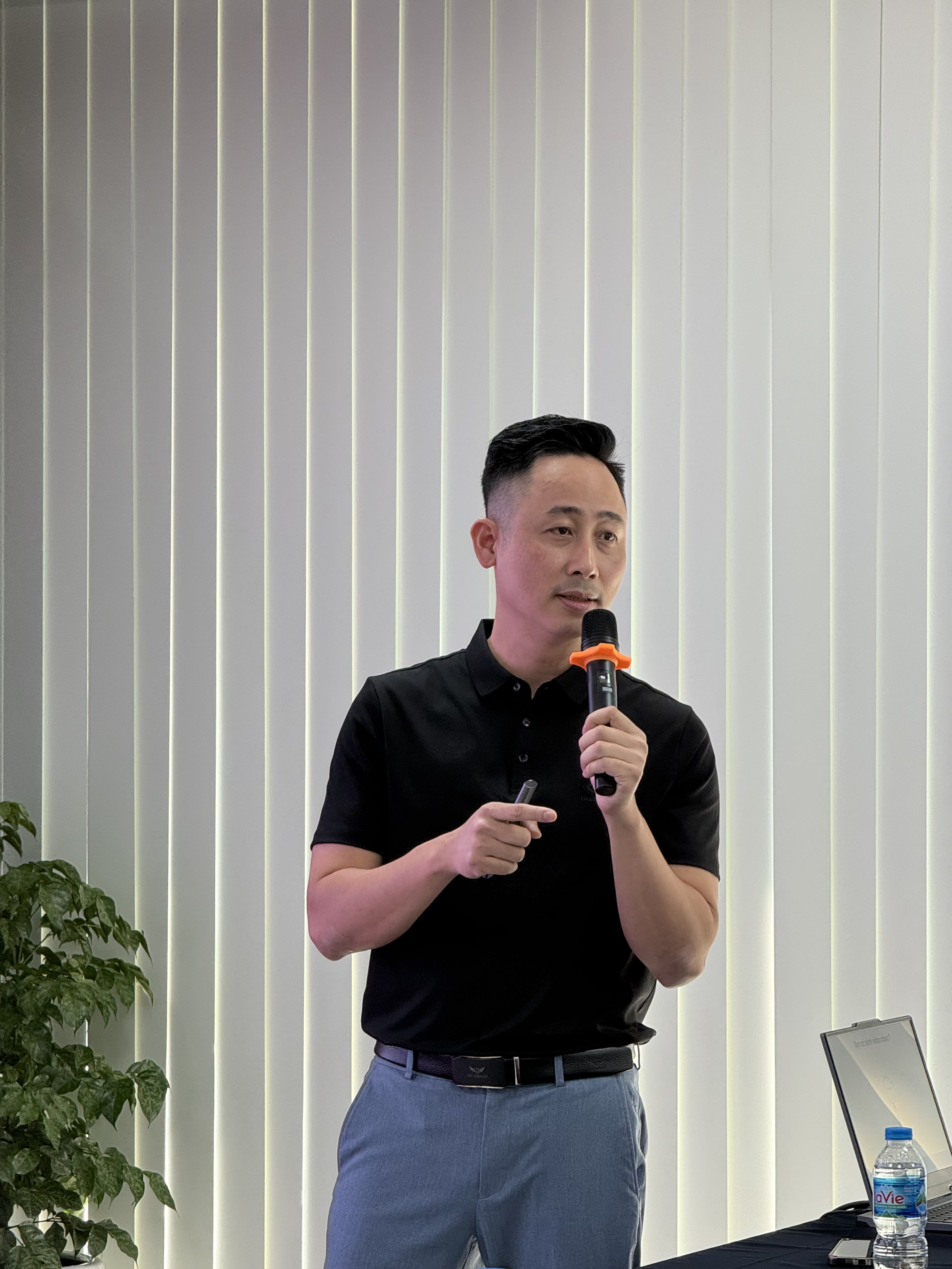 Đăng Dương áo polo đen cầm micro nói trước phông tường cây xanh trong phòng đào tạo