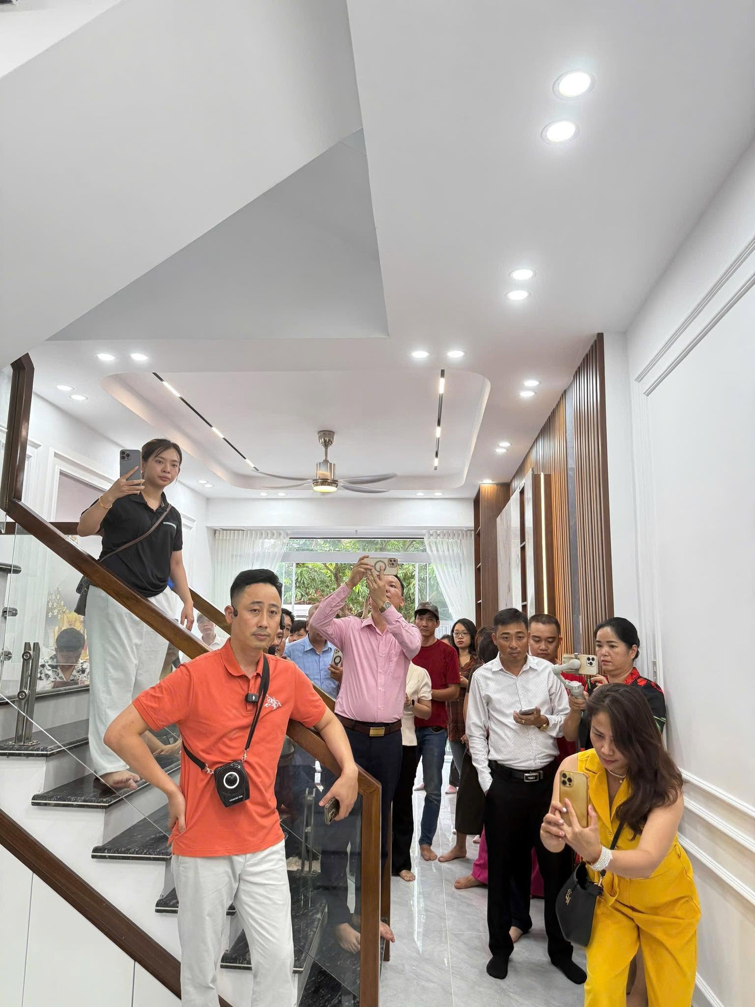 Đăng Dương mặc áo polo cam đứng trên cầu thang một căn nhà phố đã hoàn thiện, phía sau là nhóm học viên đang giơ điện thoại quay