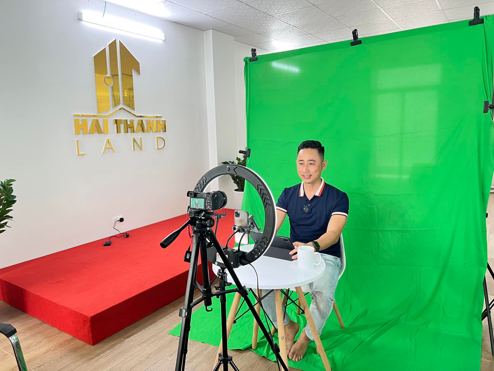 Người đàn ông ngồi trước phông xanh trong văn phòng bất động sản, có đèn tròn và máy quay đặt trên chân máy