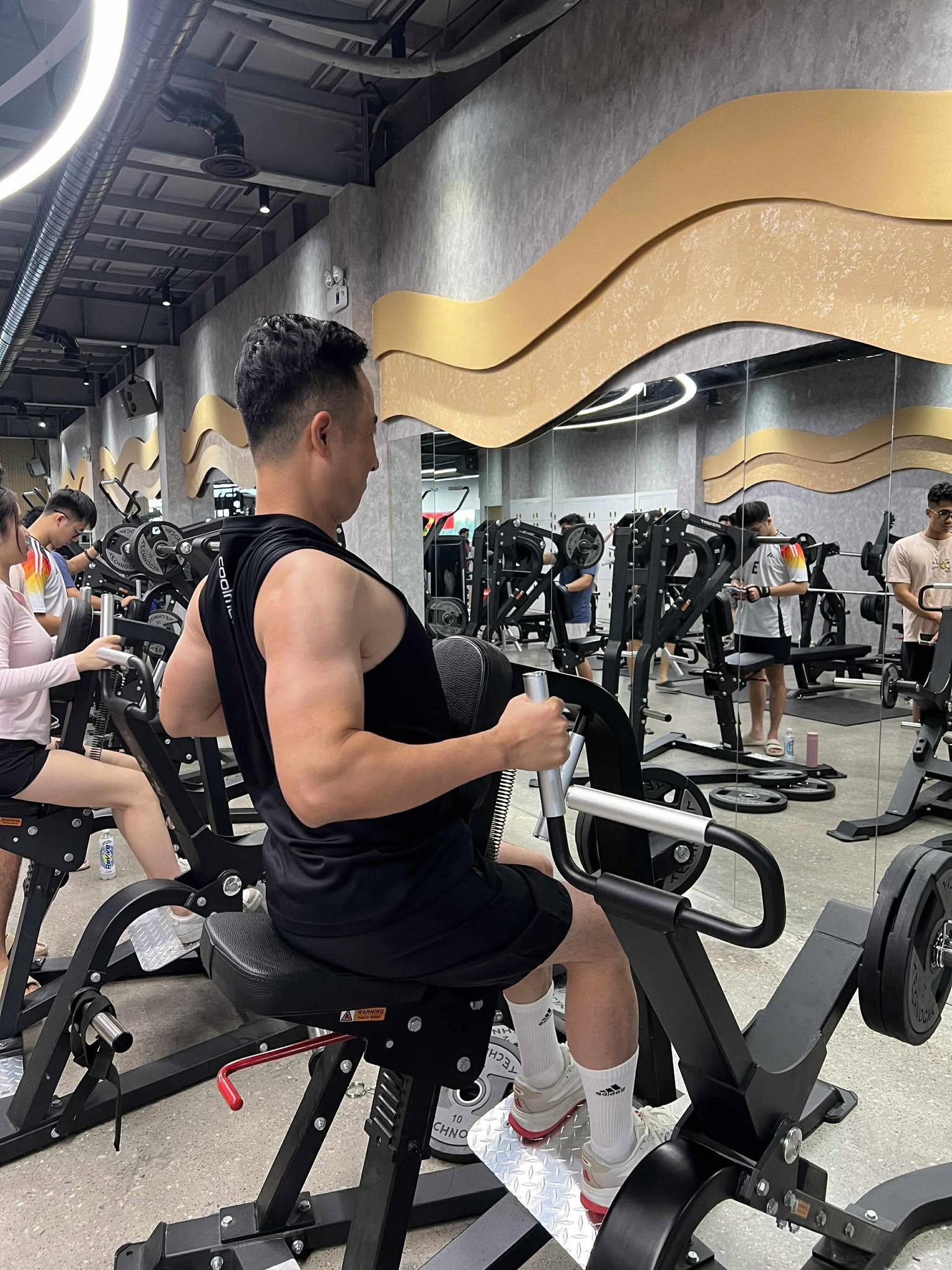 Một người mặc áo ba lỗ đen ngồi trên máy tập trong phòng gym, hai tay nắm tay cầm của máy