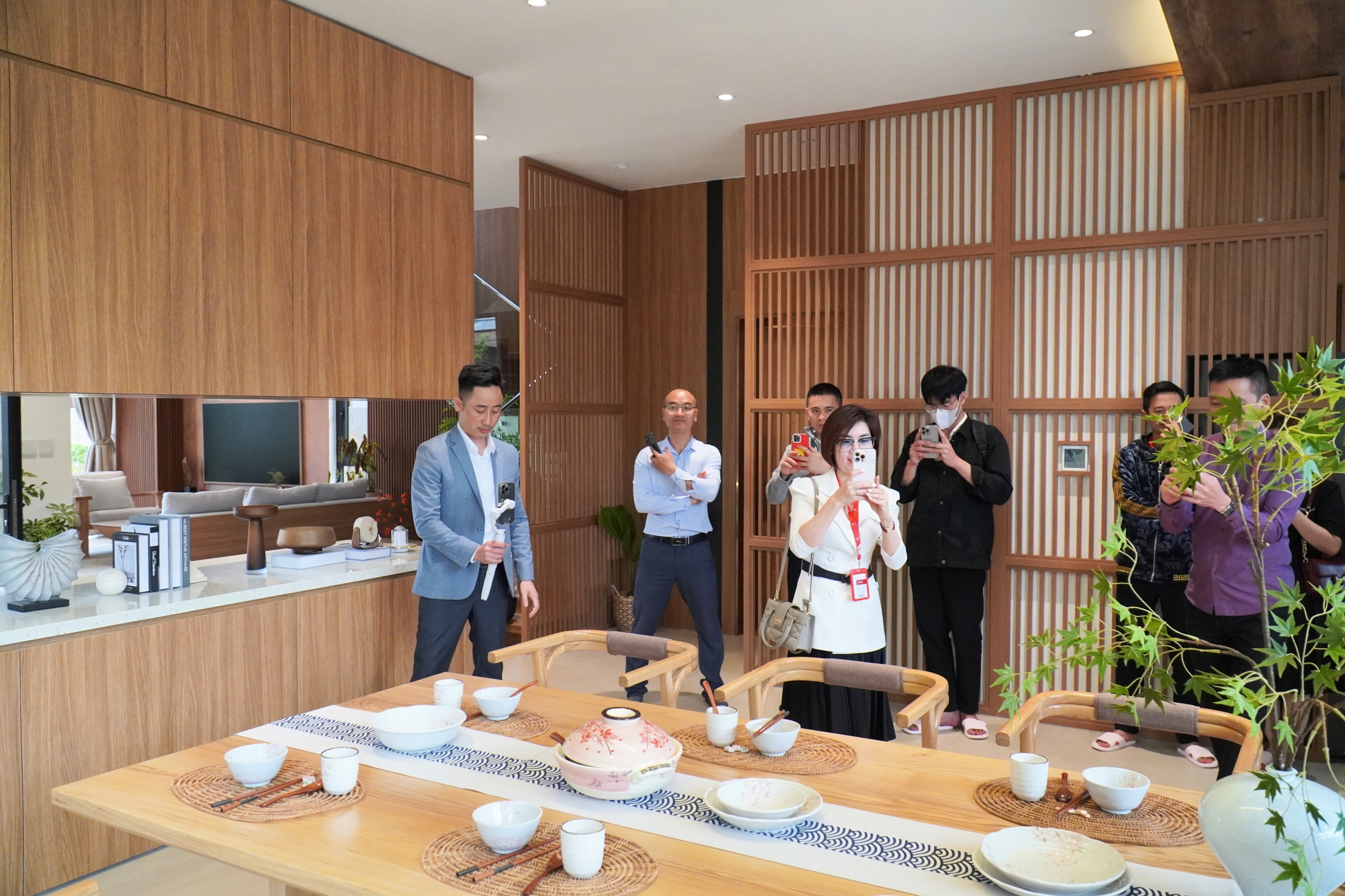 Đăng Dương mặc áo blazer xanh nhạt đứng nói và cầm điện thoại trong một căn nhà mẫu nội thất gỗ sáng, khoảng bảy người đối diện cũng giơ điện thoại quay theo