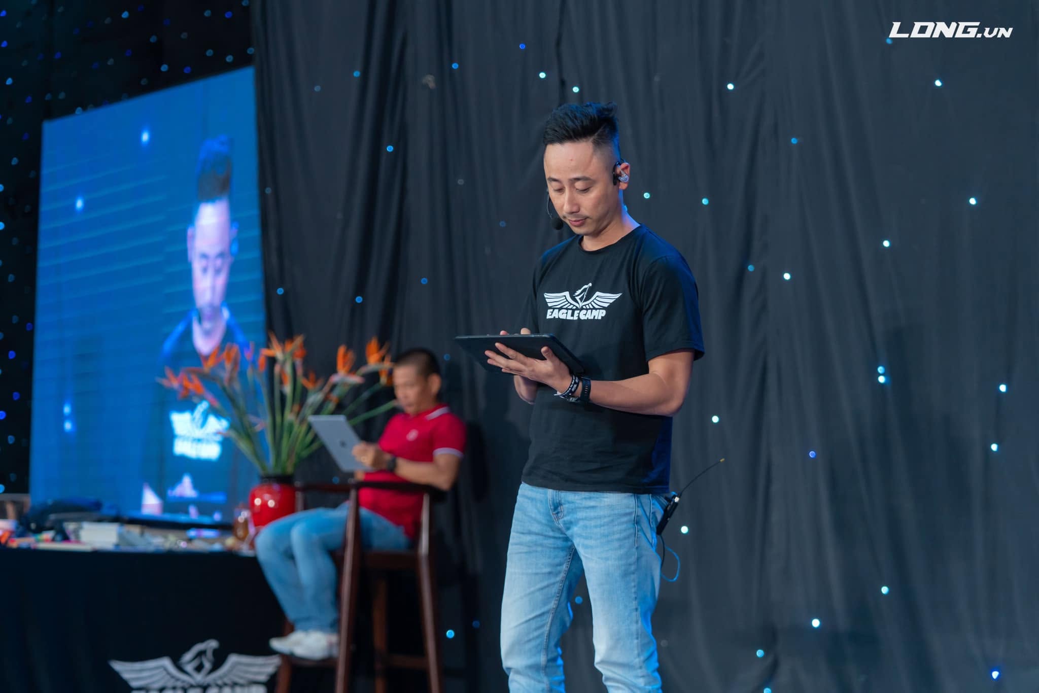 Đăng Dương mặc áo phông đen đeo micro cài đầu, cầm máy tính bảng đọc trên sân khấu có màn hình LED phía sau
