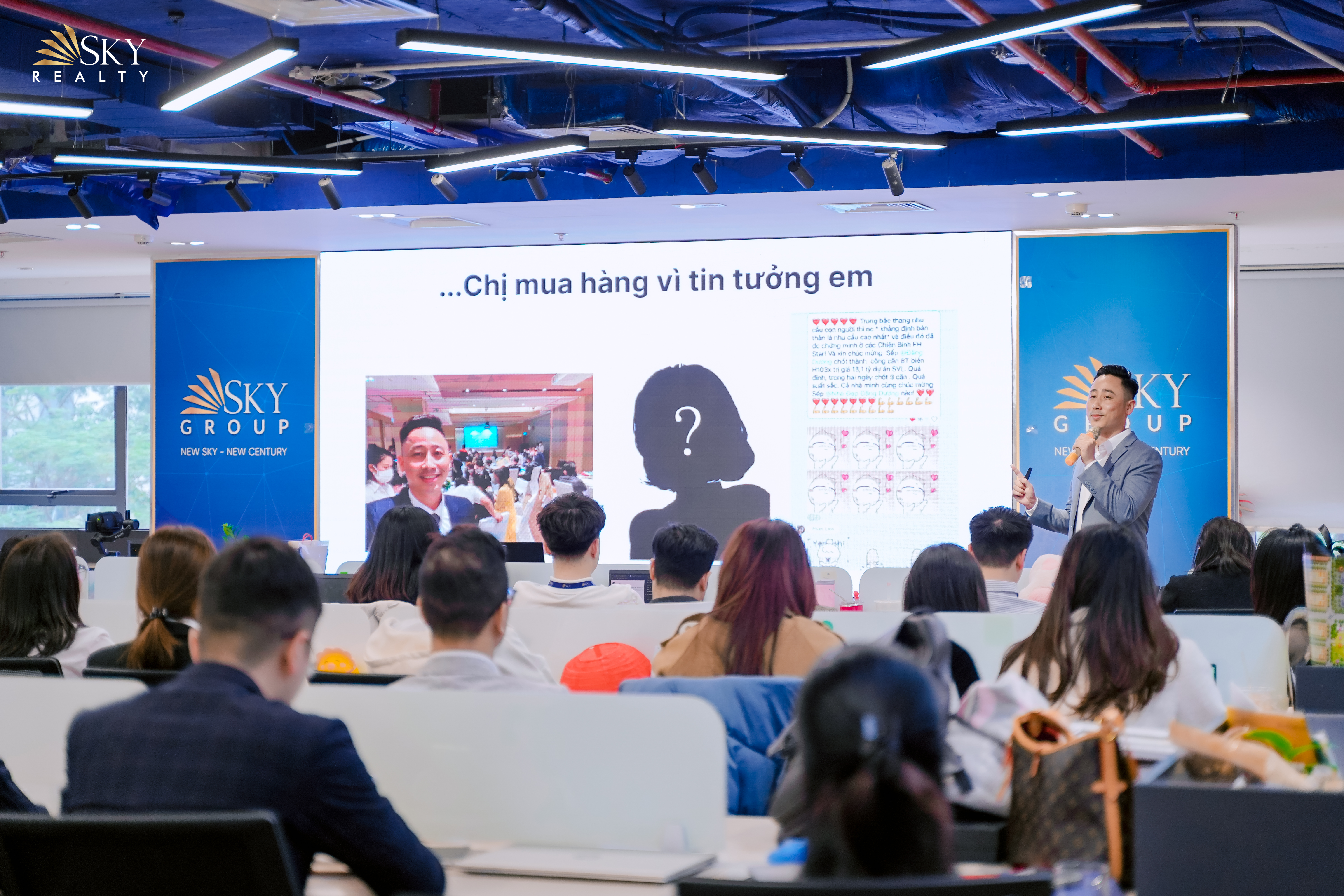 Đăng Dương mặc vest xám đứng bên phải chỉ tay về màn hình lớn, phía trước là nhiều dãy môi giới ngồi quay lưng lại máy ảnh