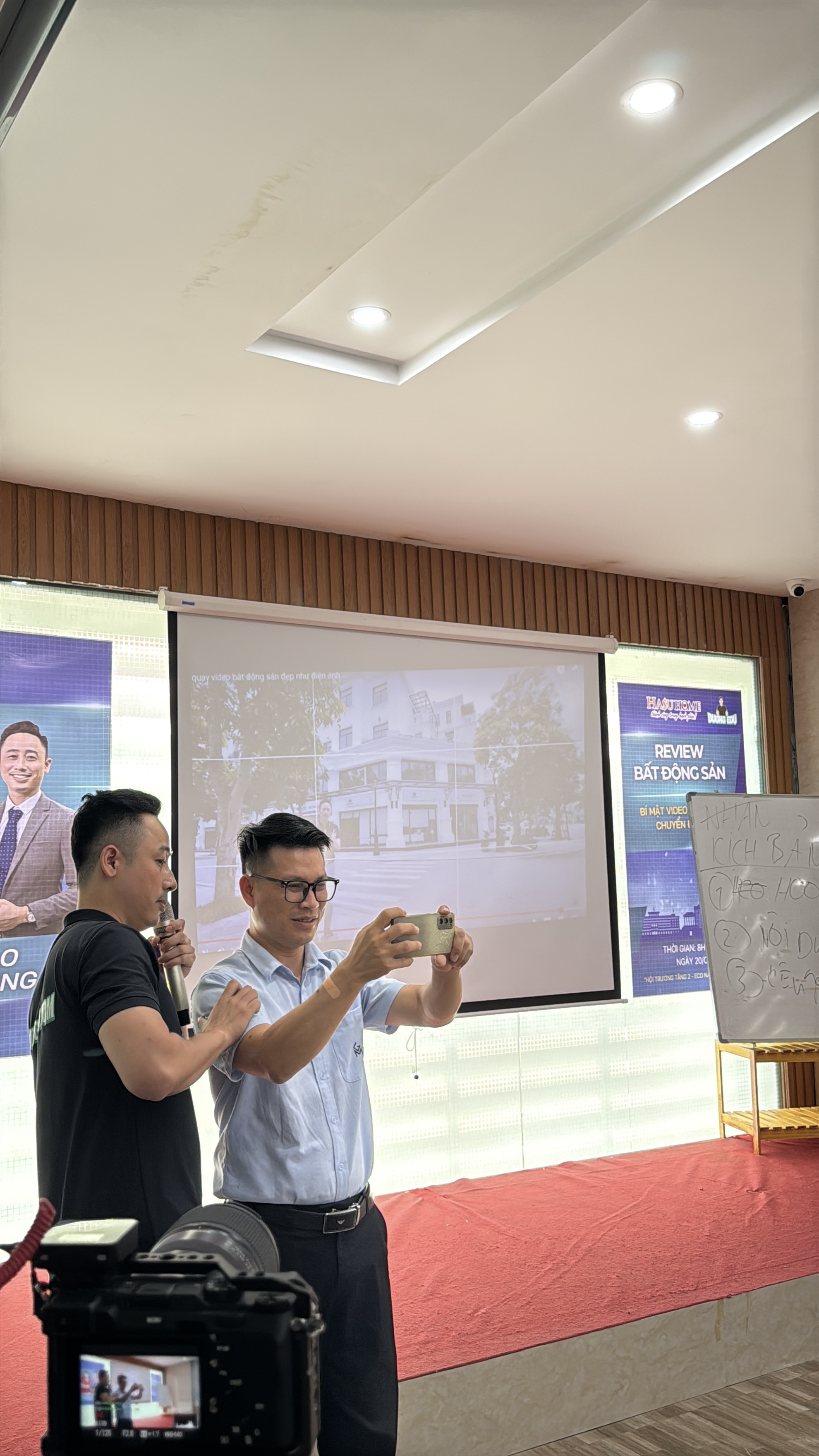 Đăng Dương mặc áo polo đen cầm micro đứng sát chỉnh lại tư thế tay cho một người đeo kính đang giơ điện thoại ngang để quay, phía sau là màn chiếu hình một căn biệt thự