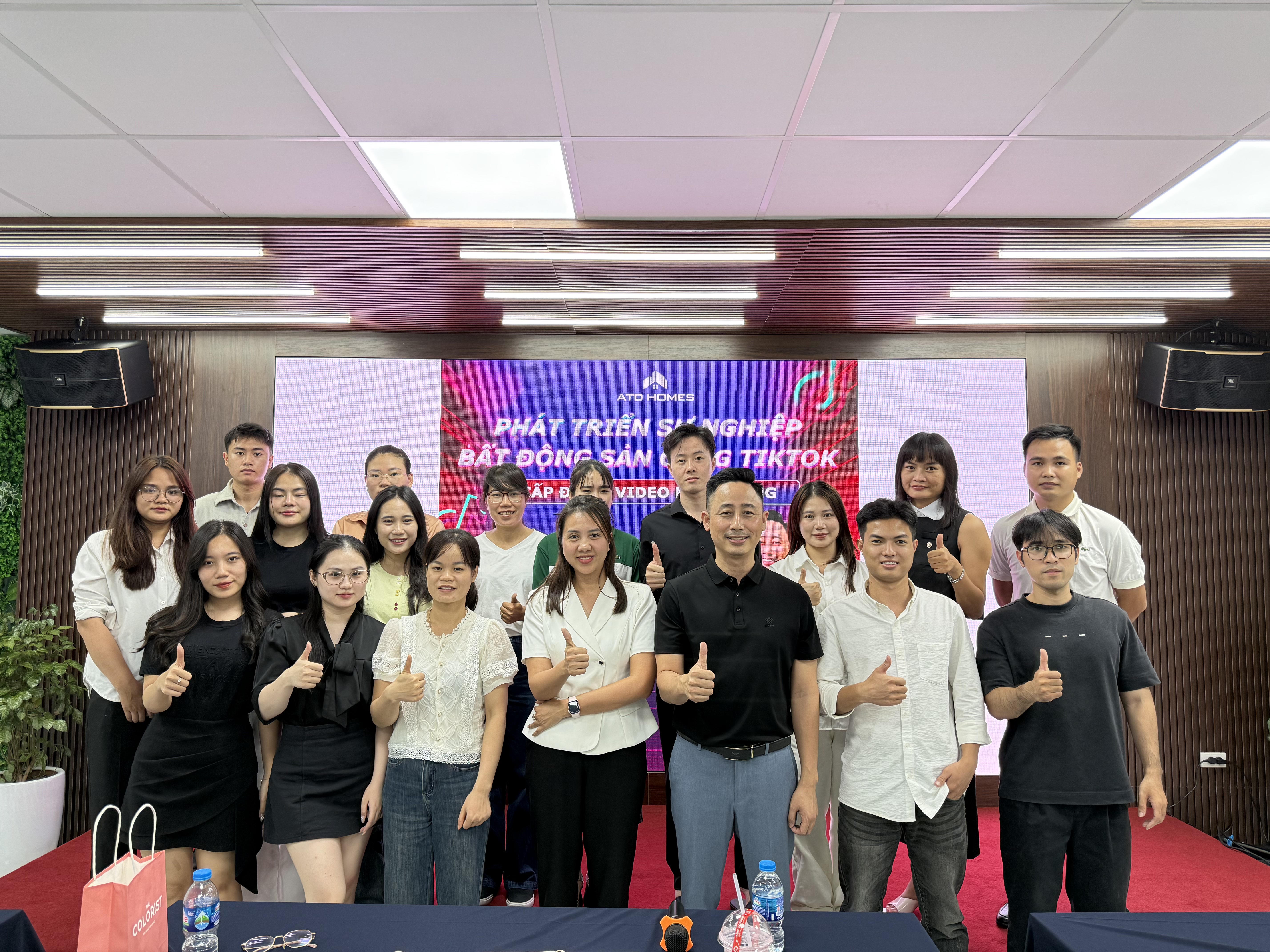 Khoảng mười tám người đứng thành hai hàng chụp ảnh chung, nhiều người giơ ngón tay cái, phía sau là màn hình lớn chạy dòng chữ về phát triển sự nghiệp bất động sản