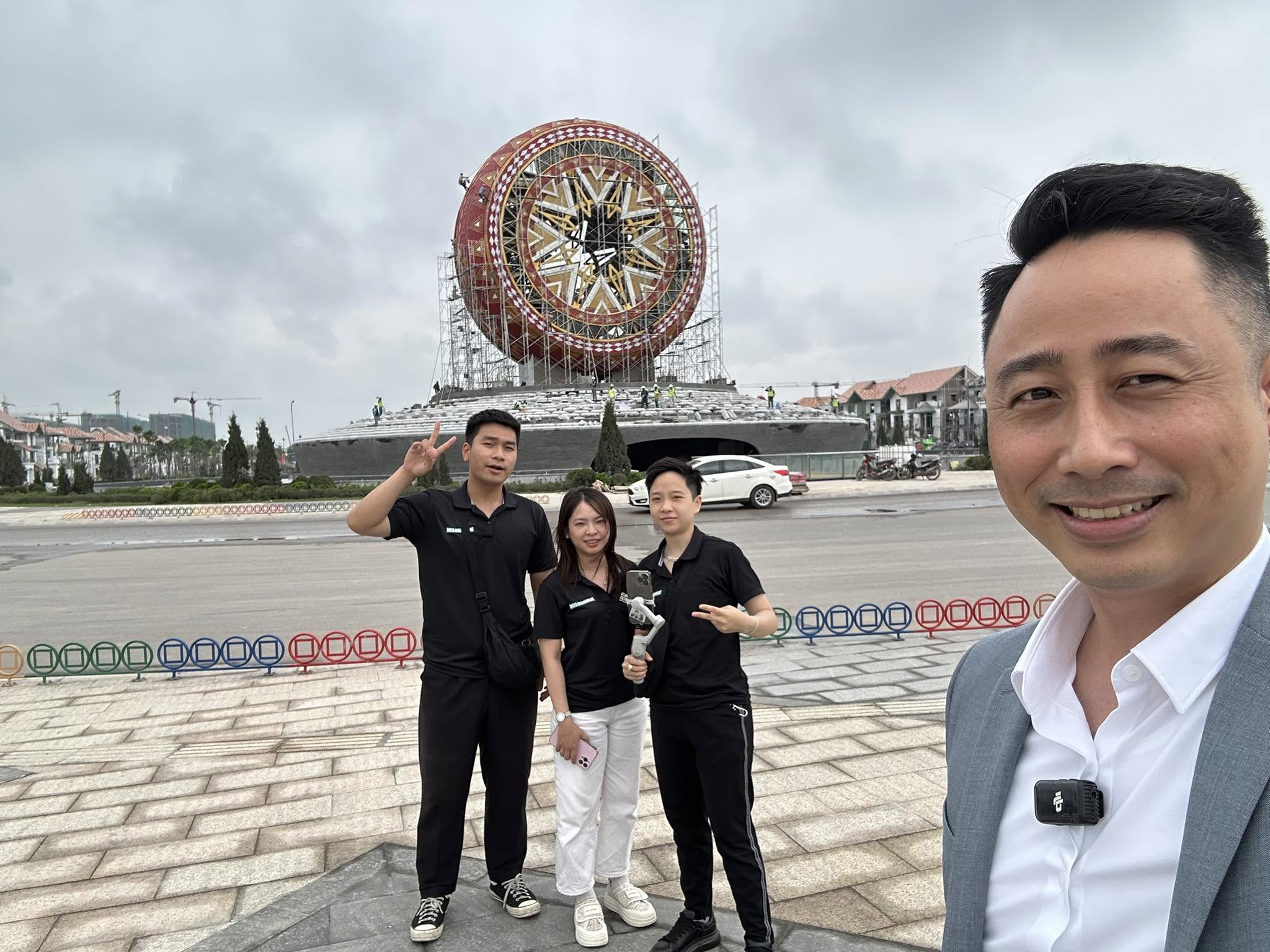 Đăng Dương chụp ảnh tự sướng cùng ba đồng nghiệp mặc áo đen cầm điện thoại gắn tay cầm chống rung tại quảng trường một khu đô thị