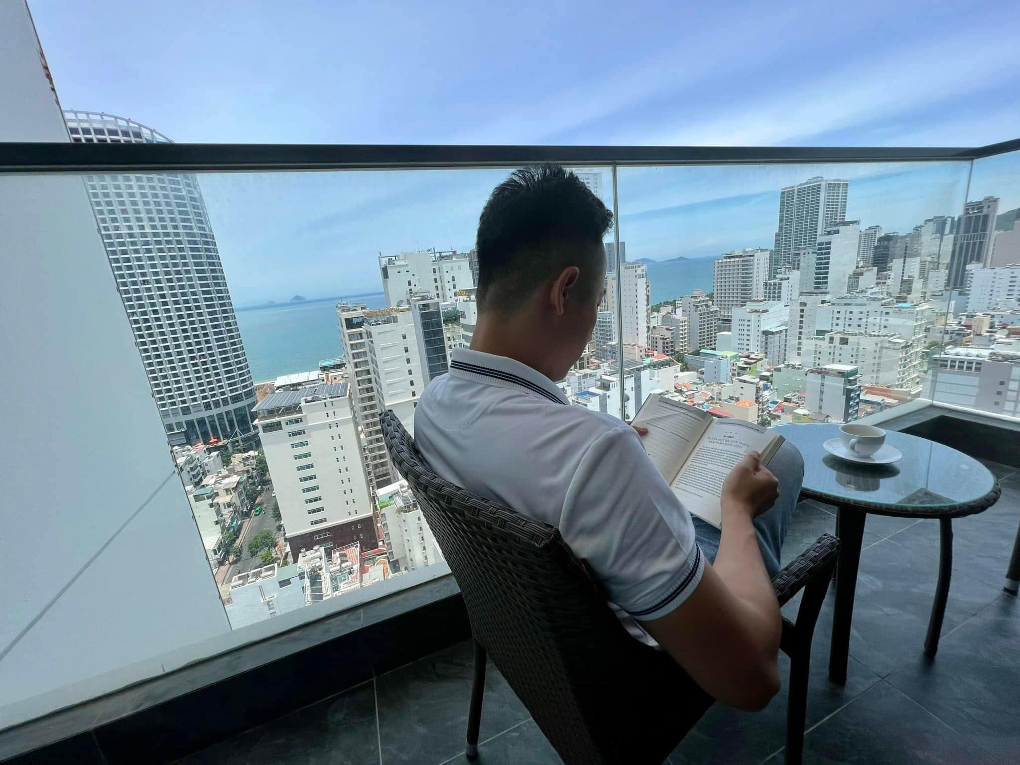 Một người ngồi trên ban công tầng cao đọc sách, bên cạnh là tách cà phê, phía dưới là thành phố ven biển