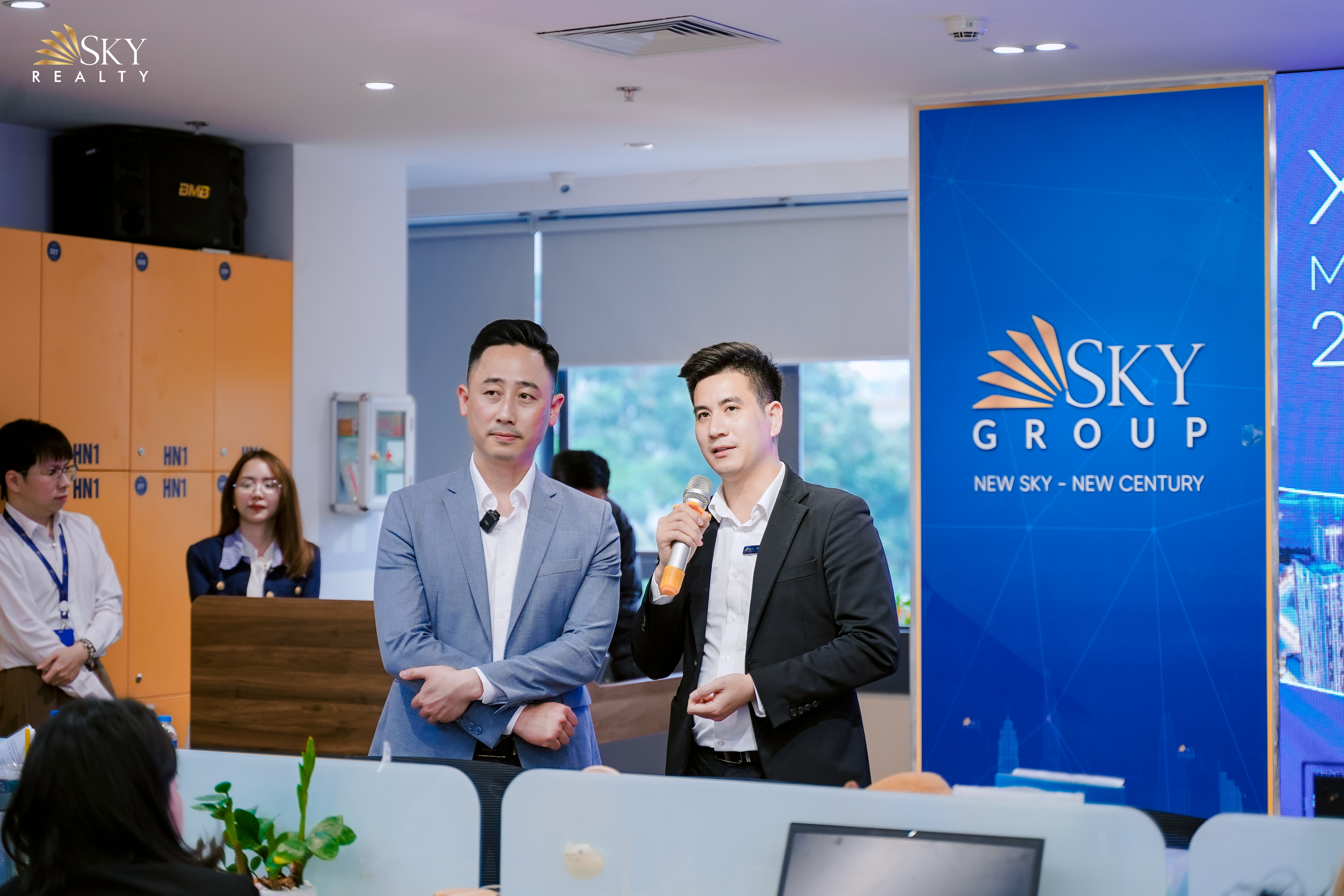 Hai người đàn ông mặc vest đứng cạnh nhau, một người cầm micro đang nói, người còn lại khoanh tay lắng nghe, phía sau là standee và tủ để đồ của văn phòng