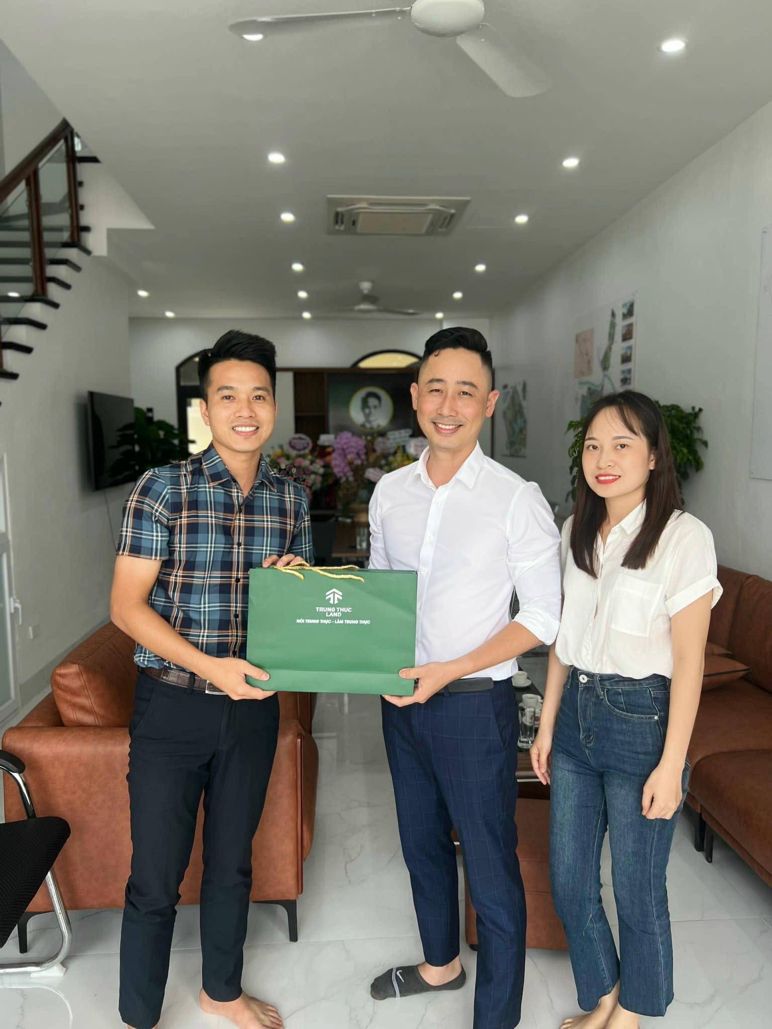 Đăng Dương mặc áo sơ mi trắng đứng giữa, cùng hai người trao nhau túi quà in chữ Trung Thực Land trong một văn phòng mới khai trương