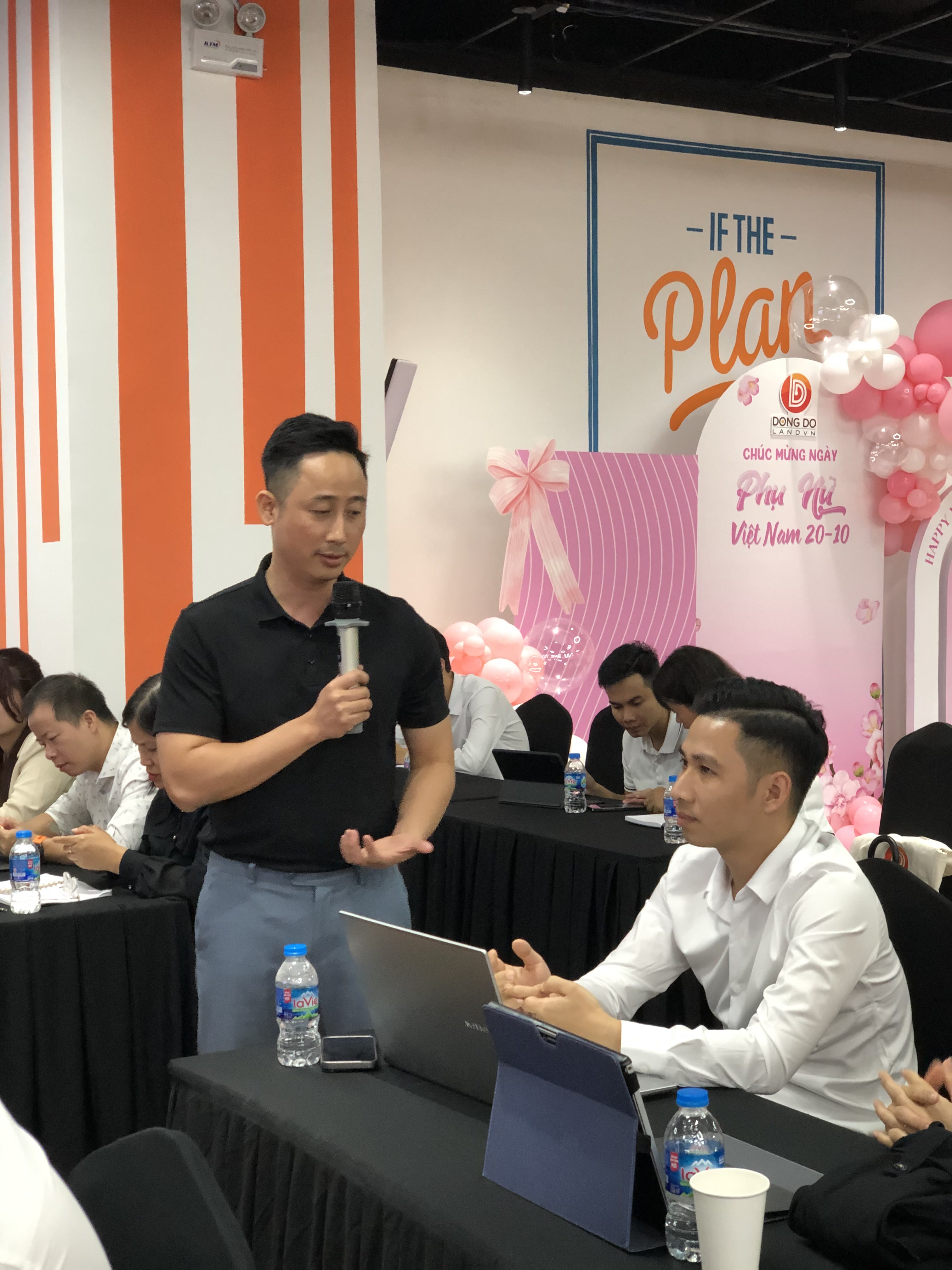 Đăng Dương mặc áo polo đen cầm micro cúi xuống trao đổi riêng với một học viên nam đang ngồi trước laptop và một máy tính bảng dựng đứng