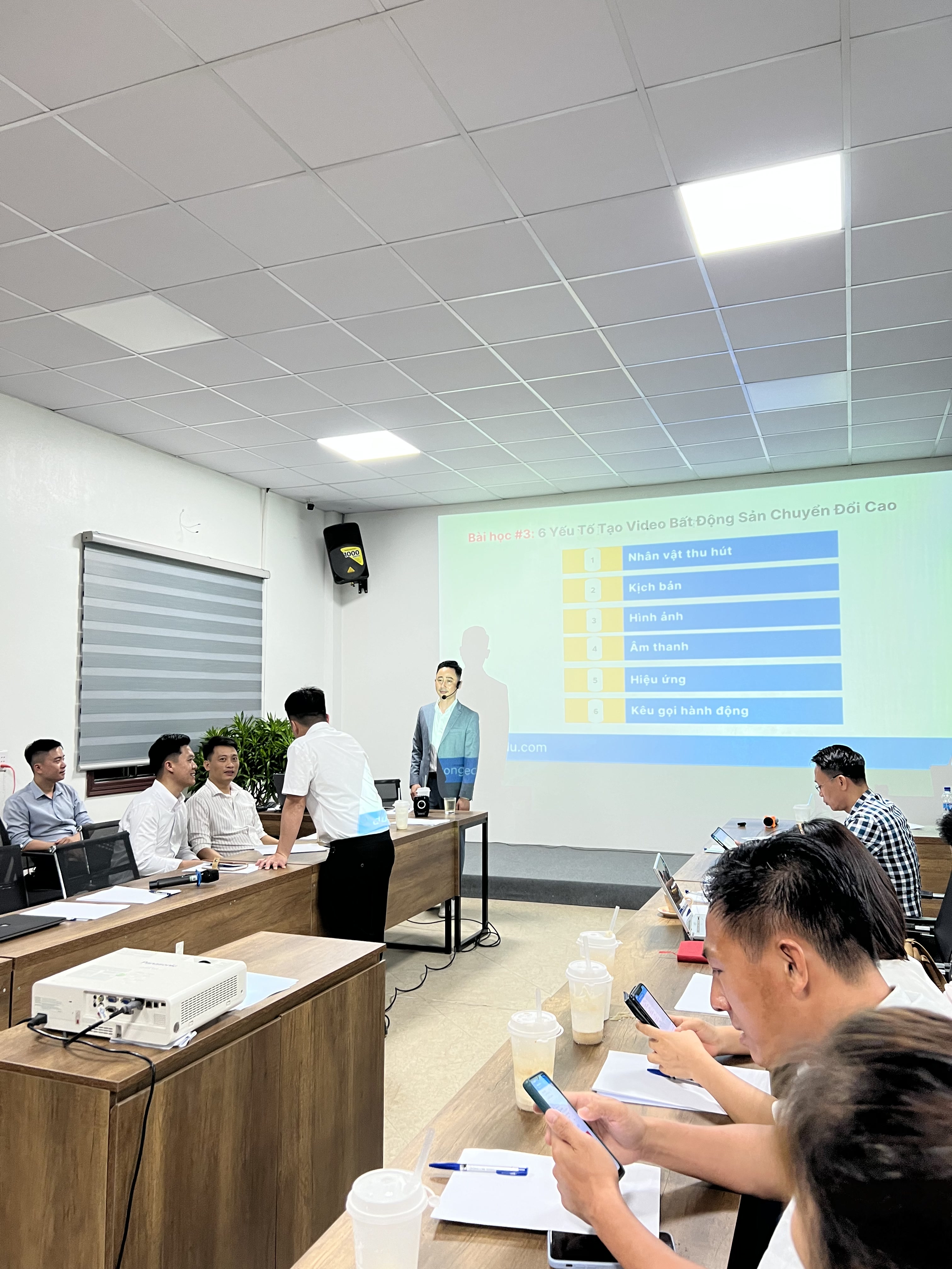 Đăng Dương mặc vest xanh xám đeo micro cài tai đứng trên bục nói, học viên ngồi quanh bàn gỗ hình chữ U với giấy bút và điện thoại trên tay