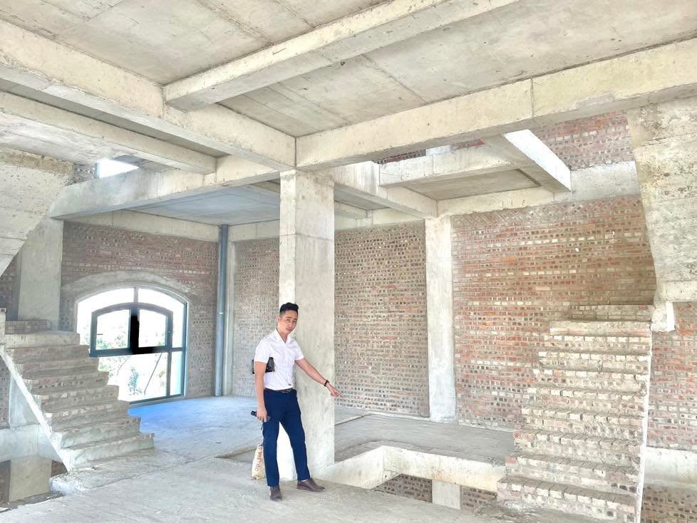 Một người mặc áo sơ mi trắng đứng bên trong căn nhà mới xây thô, tường gạch và trần bê tông chưa trát, đang chỉ tay