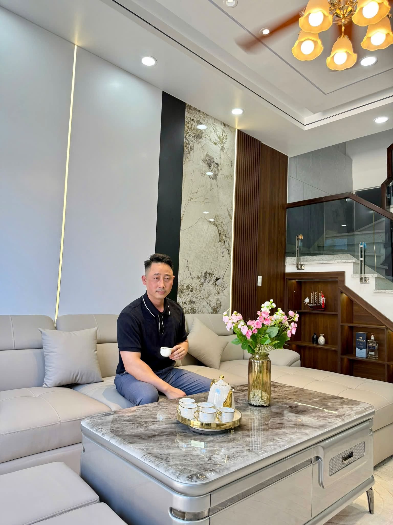 Đăng Dương ngồi trên ghế sofa trong phòng khách một căn nhà đã hoàn thiện, tay cầm chén trà, phía sau là cầu thang kính