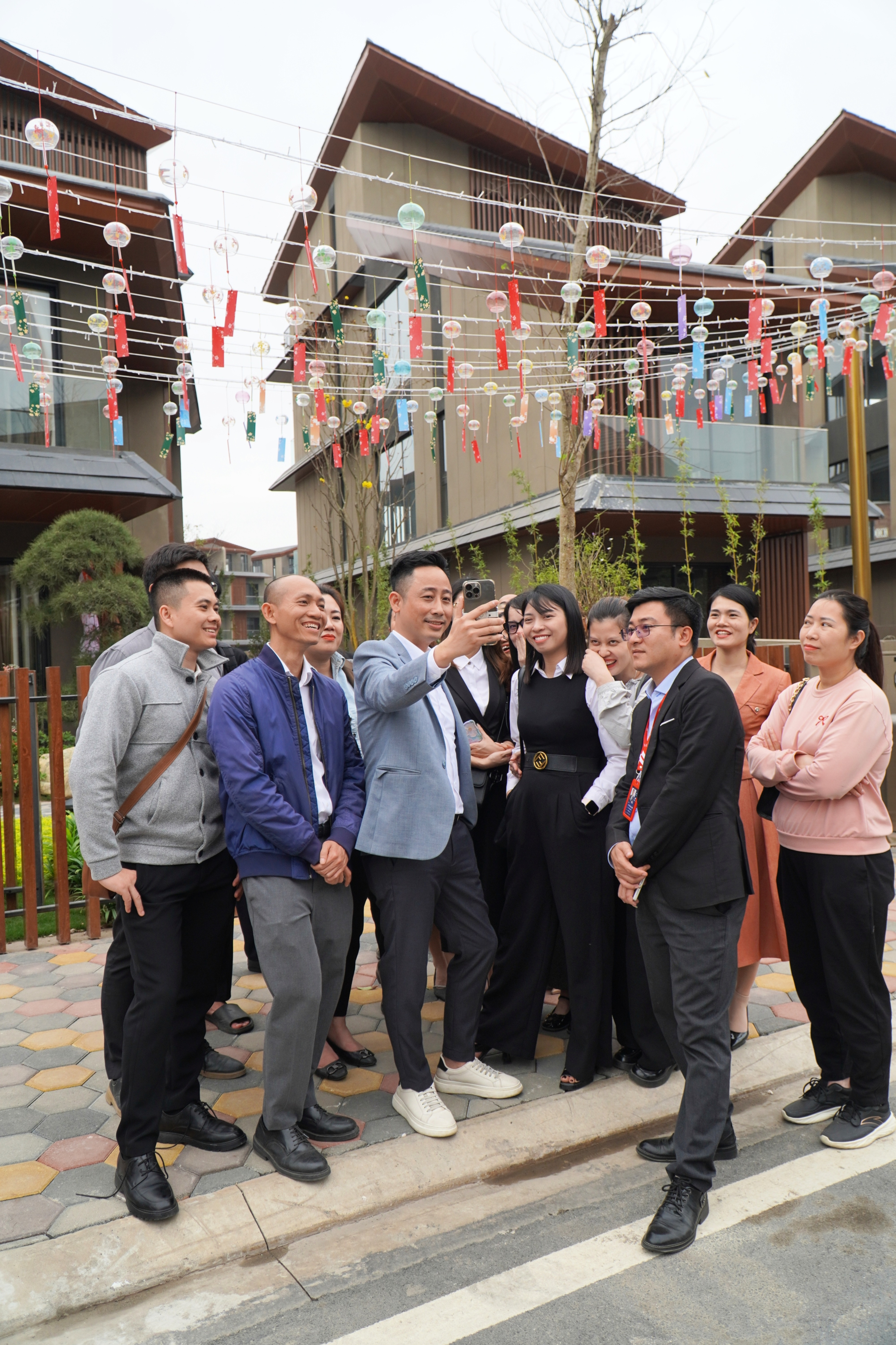 Đăng Dương mặc blazer xanh nhạt giơ điện thoại lên quay cùng khoảng mười một người vây quanh, phía trên là giàn chuông gió nhiều màu, phía sau là dãy nhà mái dốc