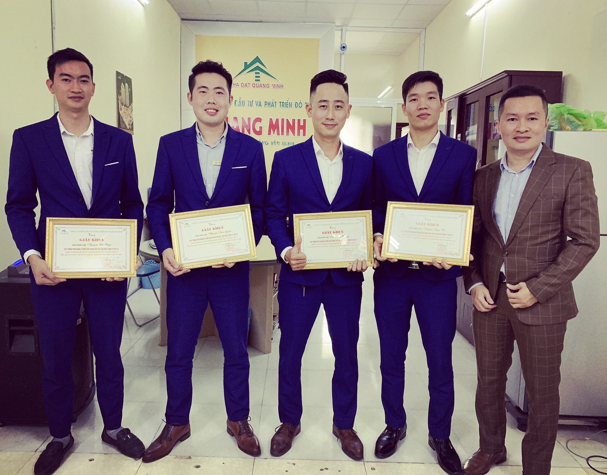 Bốn môi giới mặc vest xanh cầm giấy khen đứng cạnh một người mặc vest nâu, phía sau là biển hiệu một công ty nhà đất