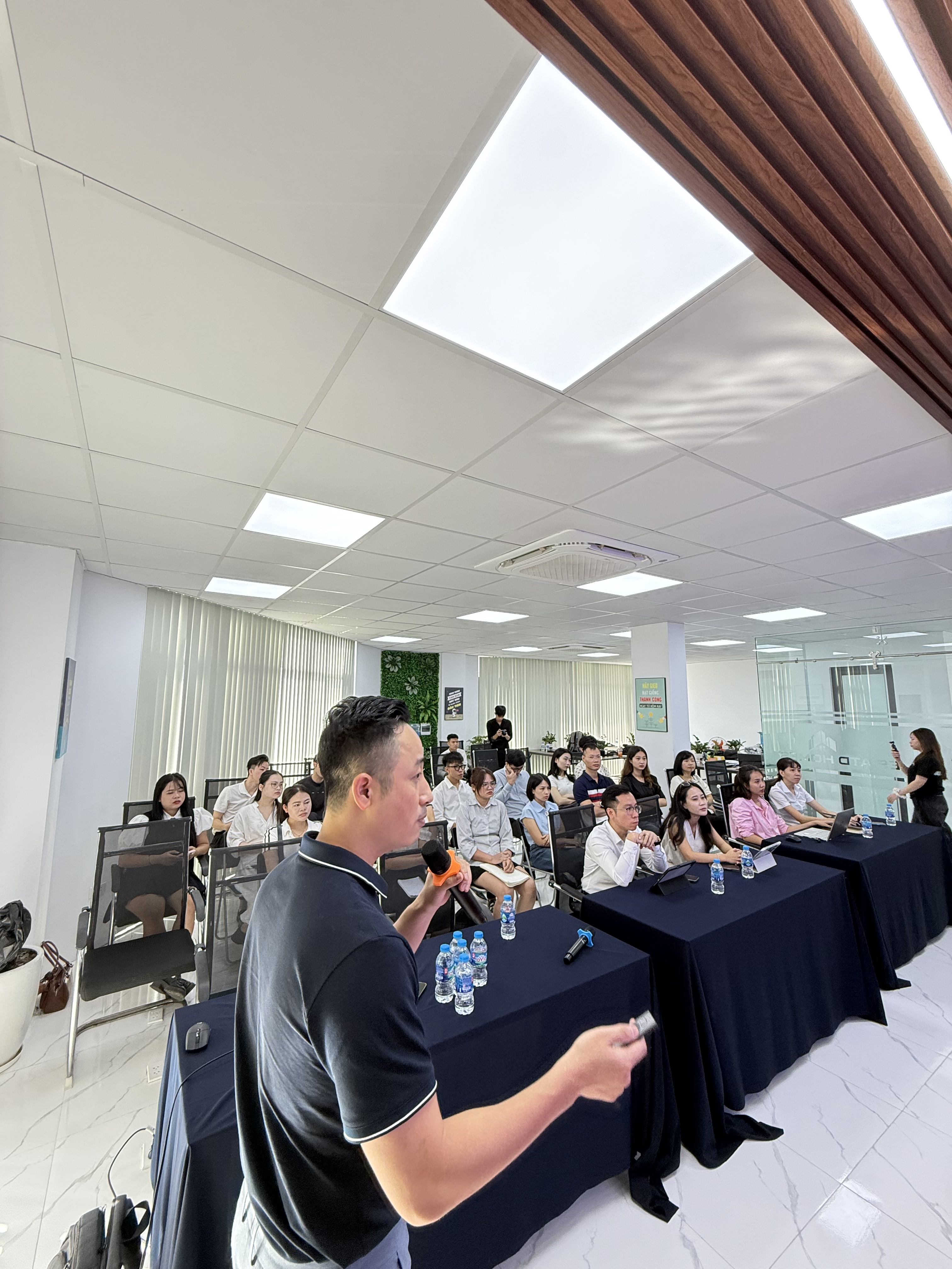 Đăng Dương đứng giữa phòng đào tạo, học viên ngồi hai bên bàn phủ khăn xanh đậm