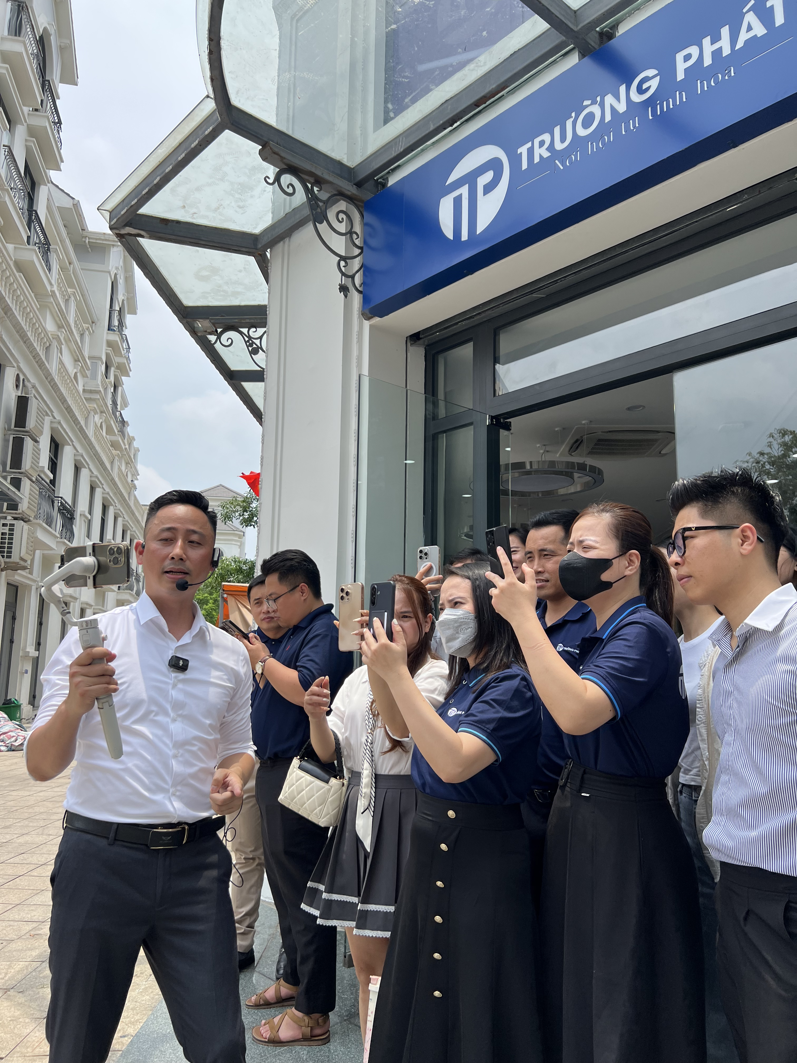 Đăng Dương mặc sơ mi trắng đeo micro cài đầu, cầm tay cầm chống rung đứng hướng dẫn trước cửa một sàn giao dịch, khoảng tám môi giới mặc áo đồng phục xanh than giơ điện thoại quay theo