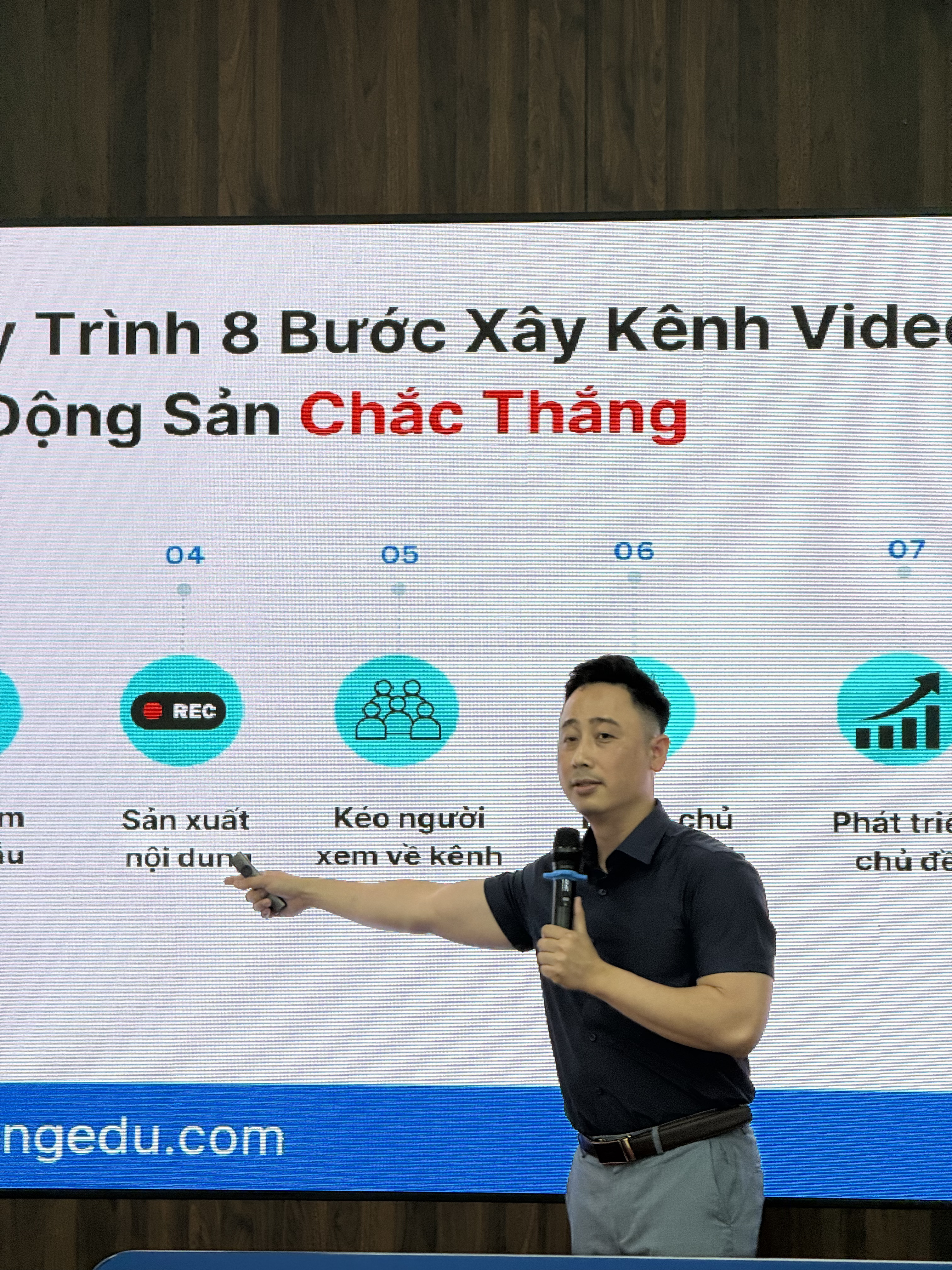Đăng Dương mặc sơ mi navy cầm micro, tay phải dùng bút trình chiếu chỉ vào màn hình đang hiện sơ đồ quy trình tám bước xây kênh video