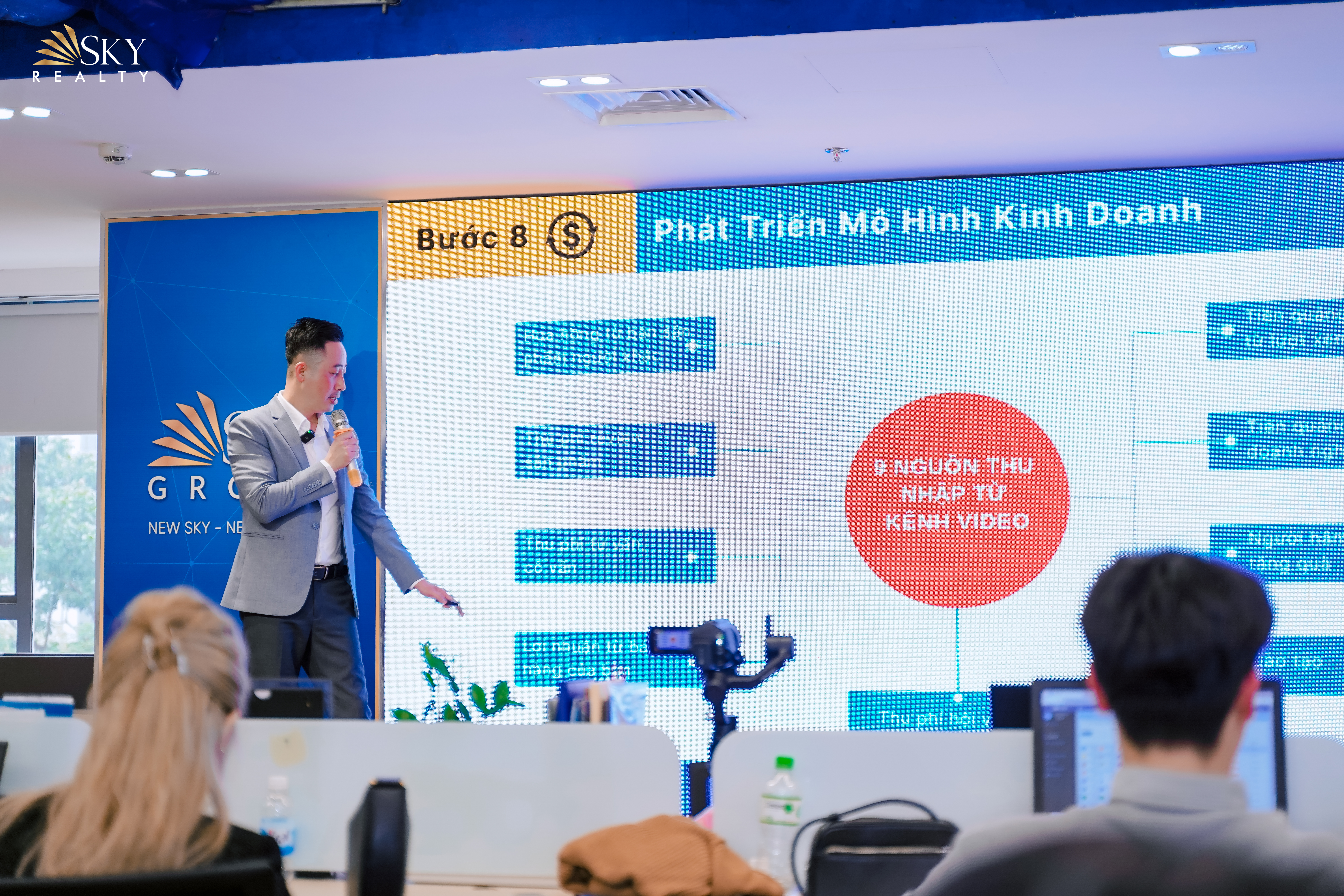 Đăng Dương mặc vest xám cầm micro, tay chỉ vào màn hình lớn đang chiếu sơ đồ các nguồn thu nhập từ kênh video, tiền cảnh là đầu hai người ngồi nghe