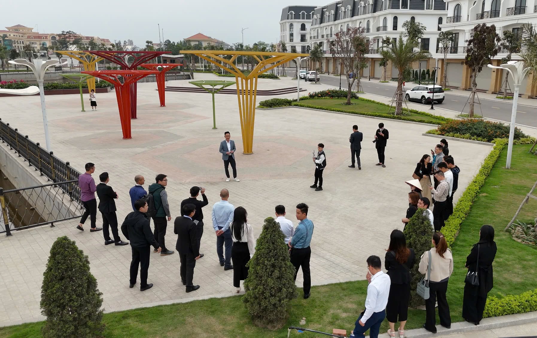 Nhìn từ trên cao, một người đứng giữa quảng trường dự án nói chuyện với khoảng hai mươi người đứng vòng quanh