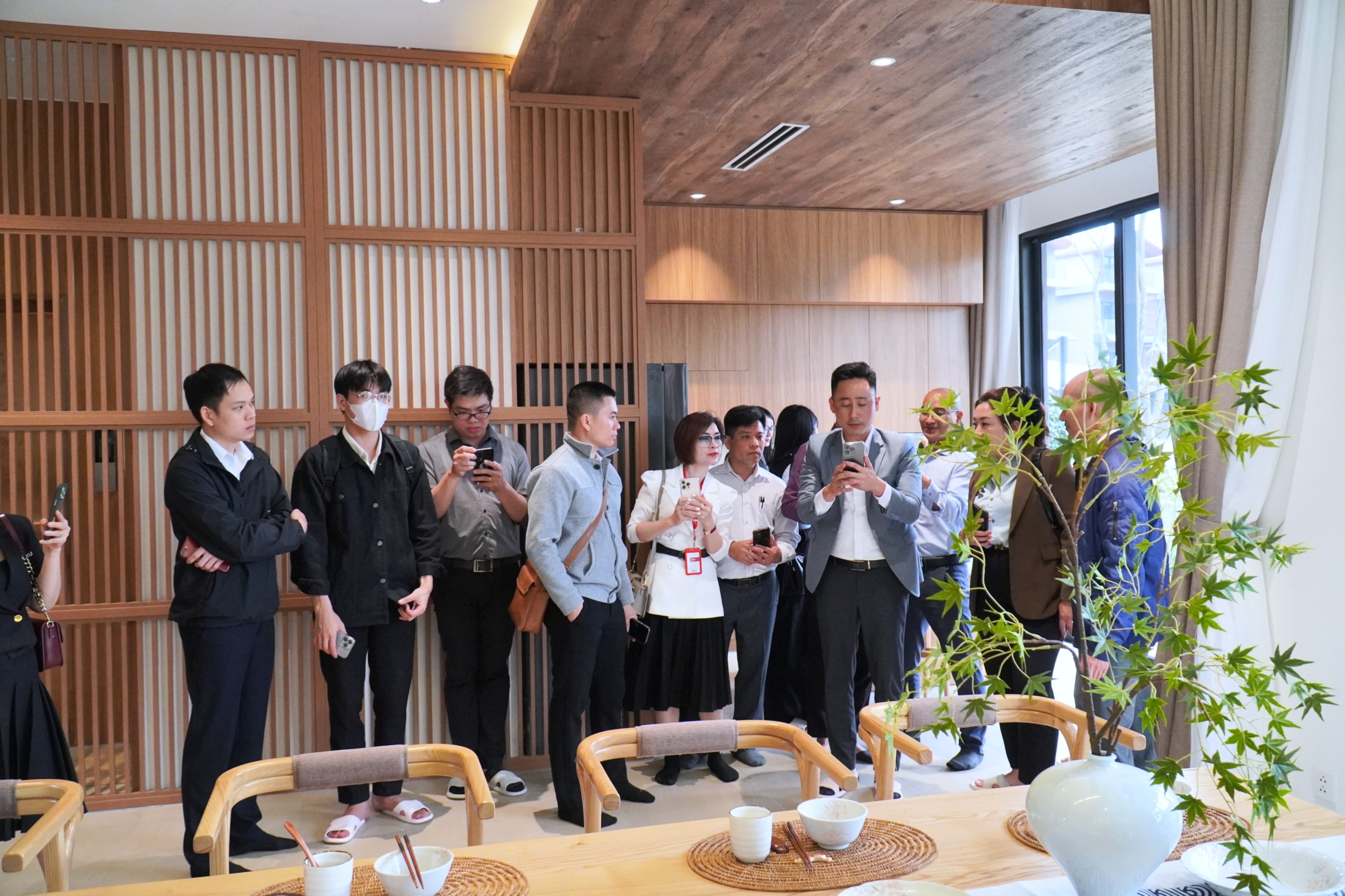 Khoảng mười hai người đứng thành hàng dọc vách gỗ trong một căn nhà mẫu, năm sáu người đang giơ điện thoại quay, tiền cảnh là bàn ăn gỗ và một cành lá xanh