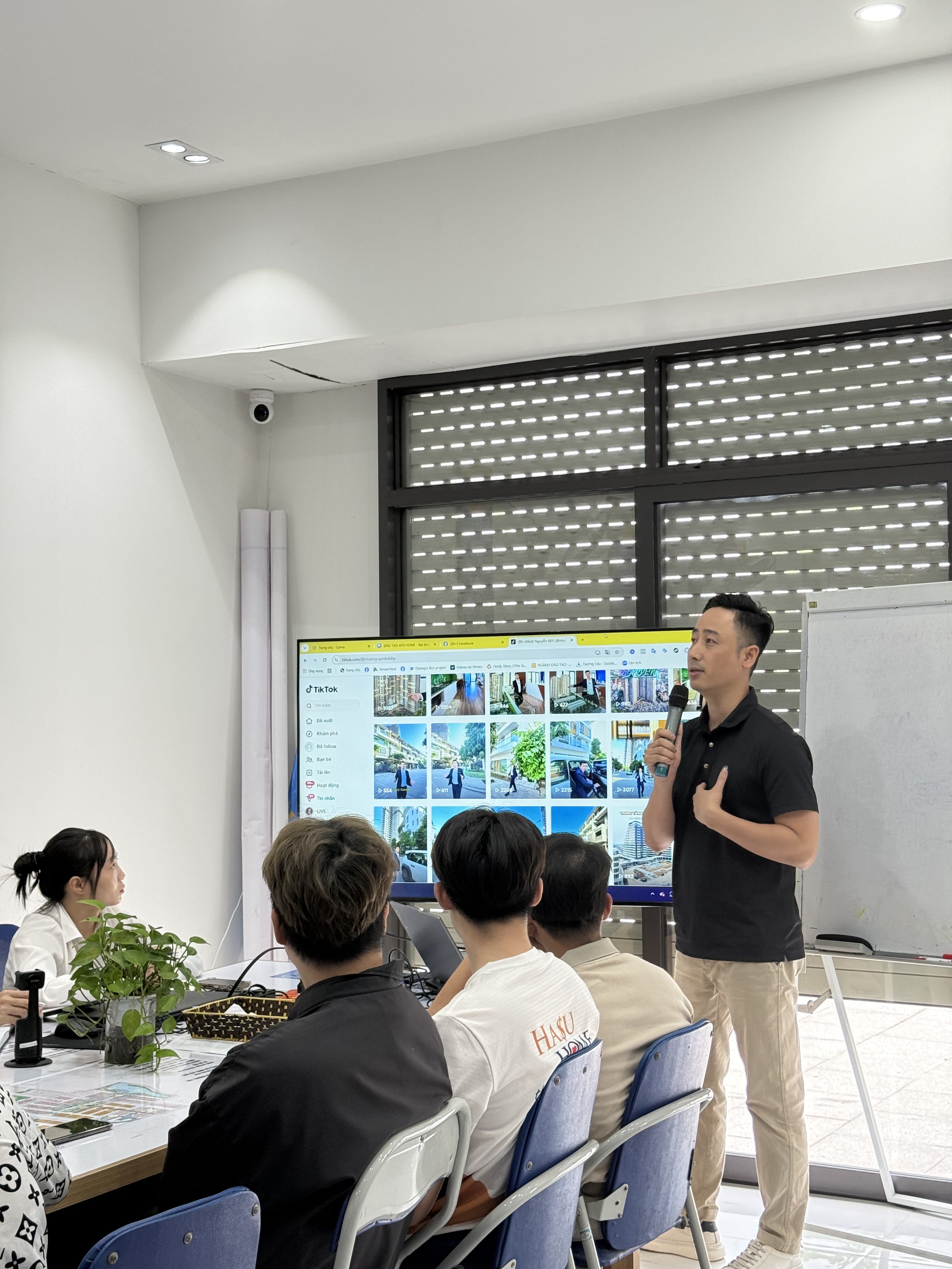 Đăng Dương mặc áo polo đen cầm micro đứng cạnh một màn hình lớn đang mở lưới video bất động sản, phía trước là mấy môi giới ngồi quay lưng theo dõi
