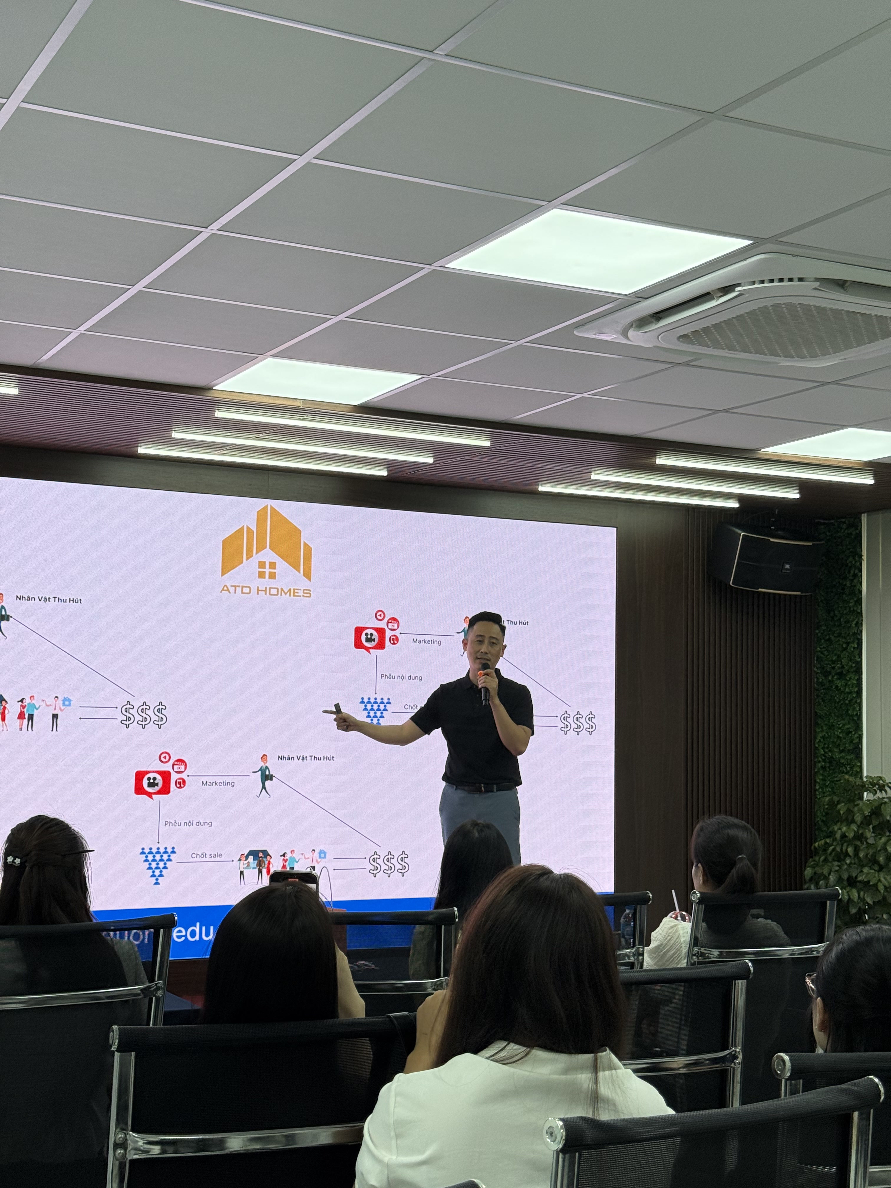 Đăng Dương mặc áo polo đen đứng bên phải một màn hình LED lớn, tay chỉ vào sơ đồ phễu đi từ nhân vật thu hút qua nội dung tới bước chốt bán hàng